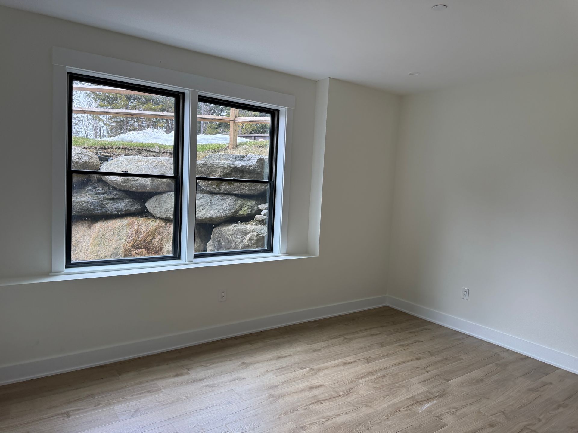 A bright, empty room with light wood floors, off-white walls, and a large window looking out onto a stone retaining wall.