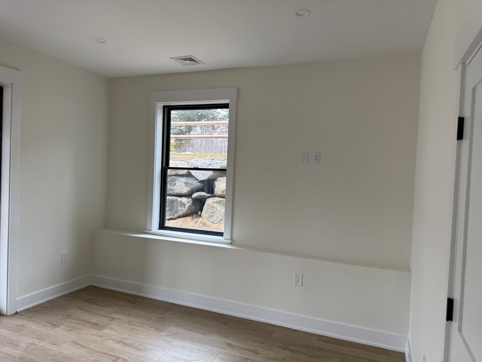 An empty room with light wood flooring, beige walls, a single window, and a built-in ledge under the window frame.