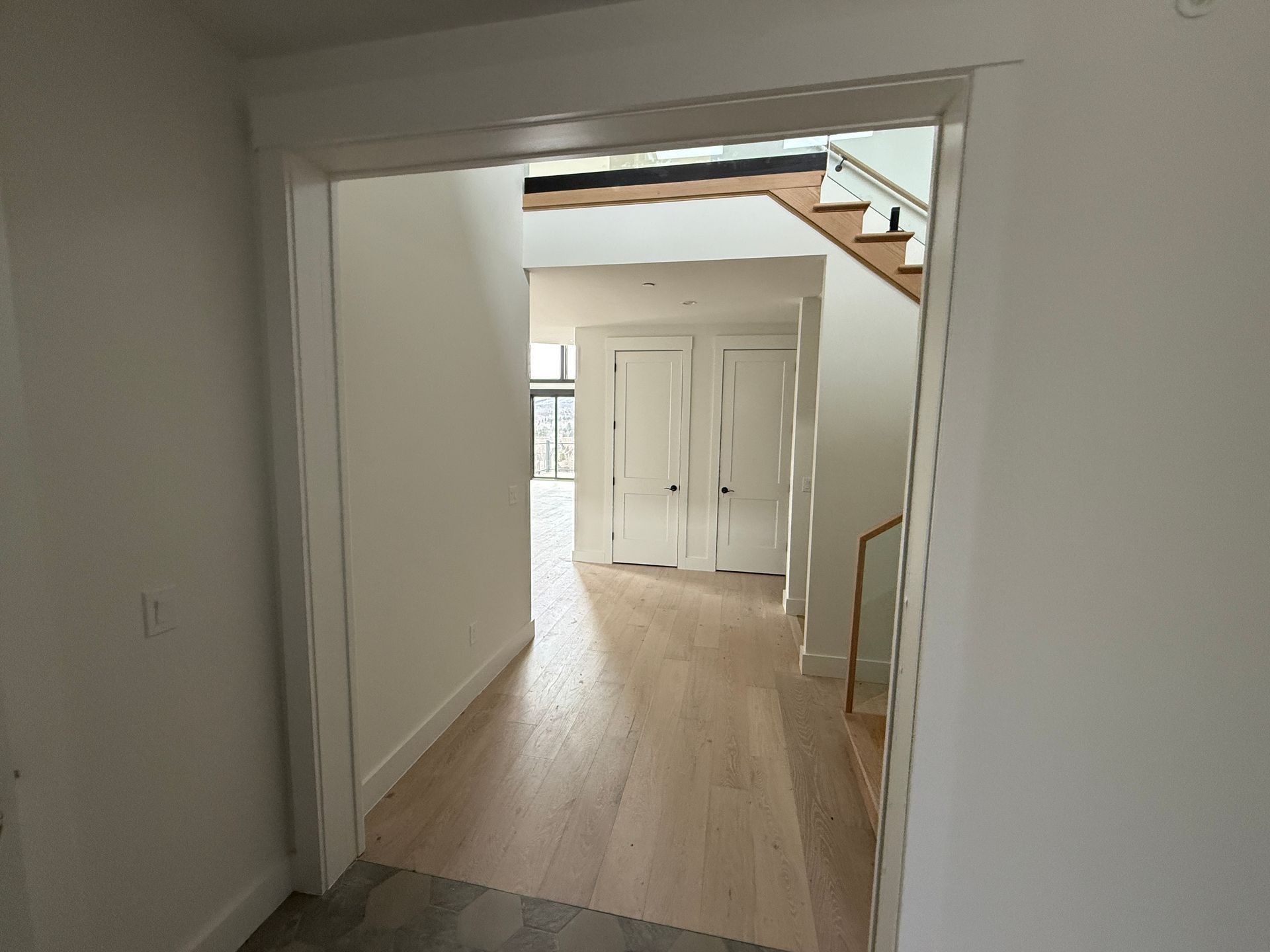 A view through a white doorway into a bright hallway with light wood floors, white walls, and a staircase.