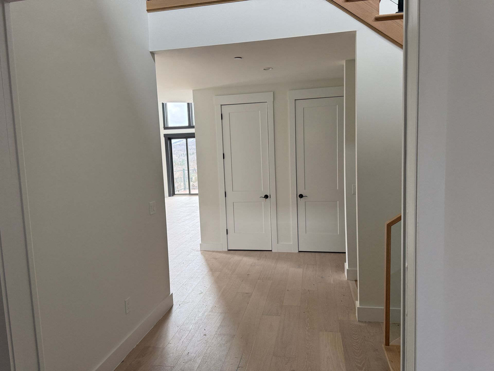 A view down a hallway with light hardwood floors leading to two white doors and a glimpse of a windowed room beyond.