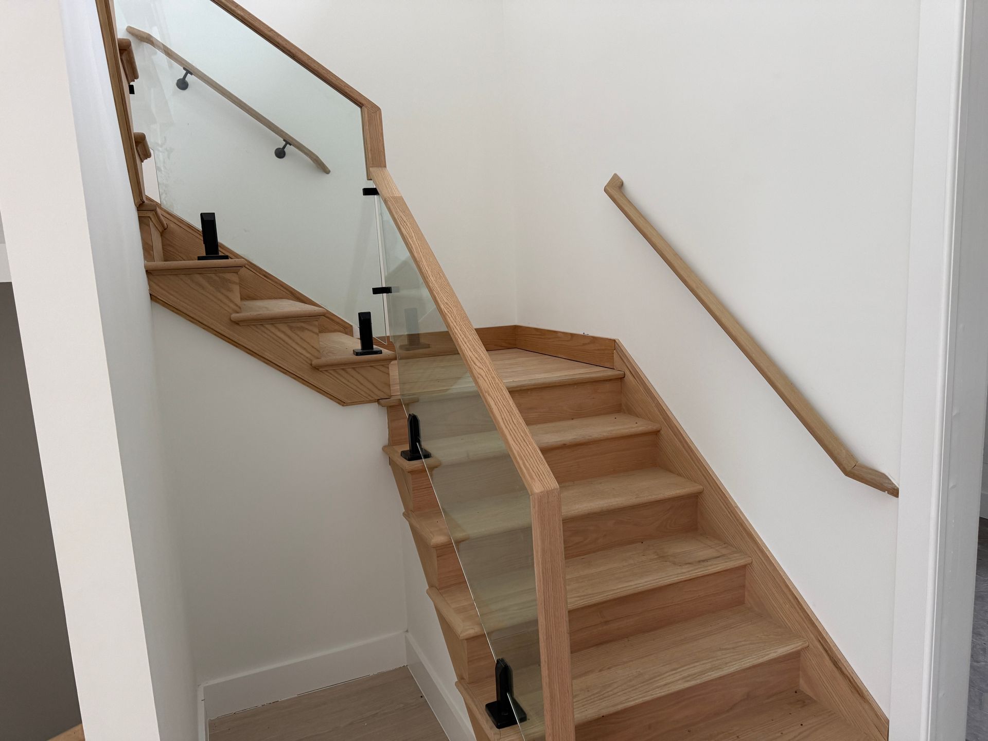 A staircase with light wood steps, a glass railing with a wood handrail, and a matching wood handrail on the white wall.