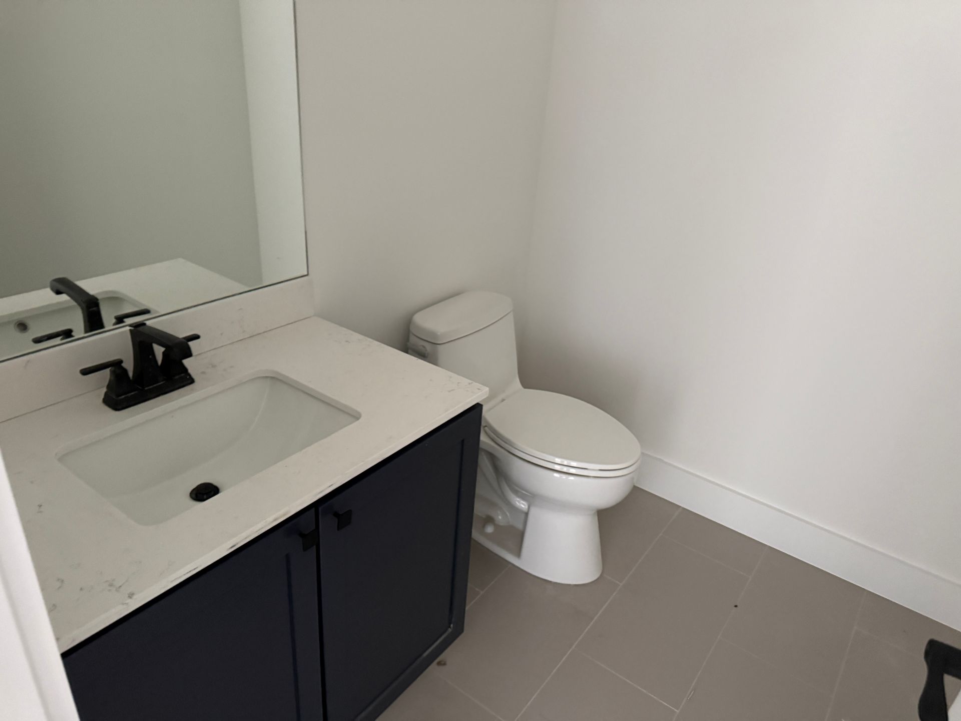 A modern bathroom featuring a white vanity with a black faucet, a white countertop, and a white toilet on gray tile.