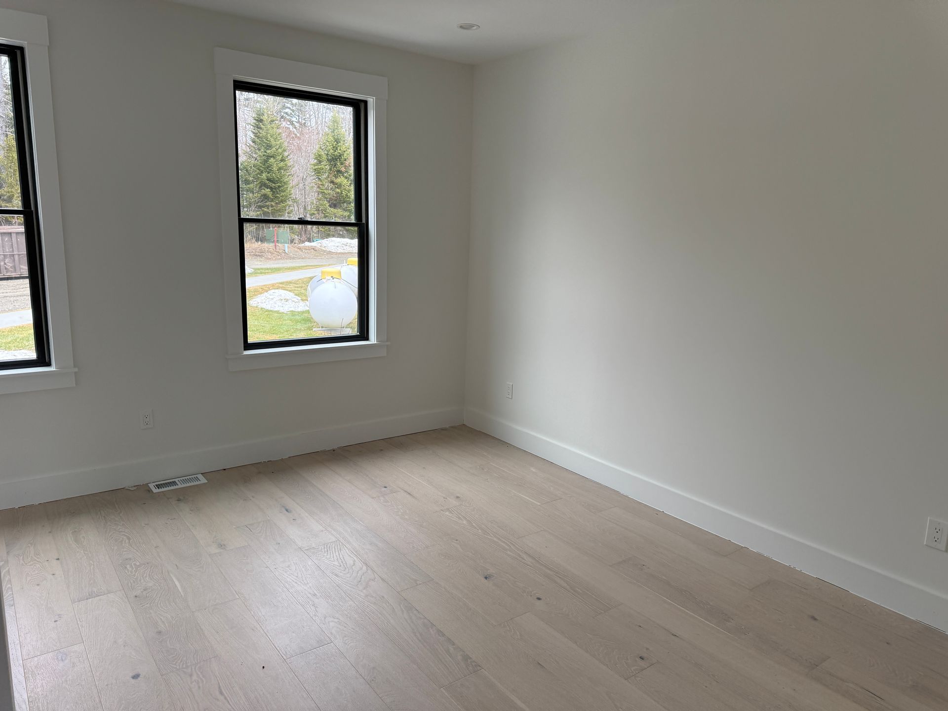 A bright, empty room with light wood-tone flooring, white walls, and two windows looking out onto a grassy area.