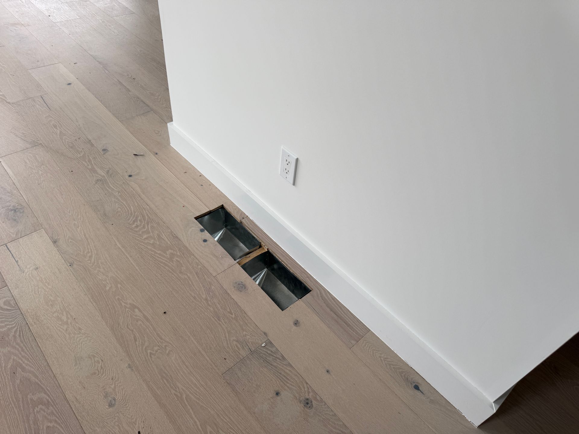 An open HVAC floor vent cut into light-colored hardwood flooring next to a white wall with an electrical outlet.