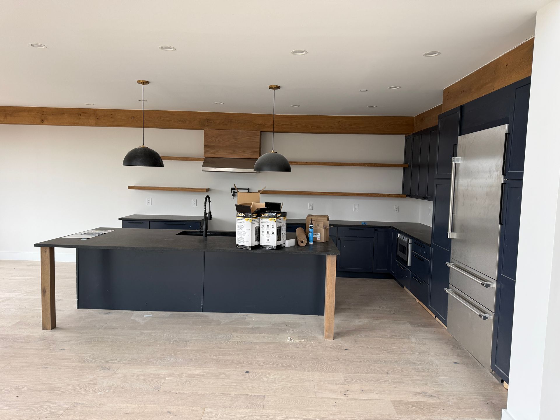 A modern kitchen with a black island, matching navy cabinetry, stainless steel refrigerator, and wood shelving and beams.