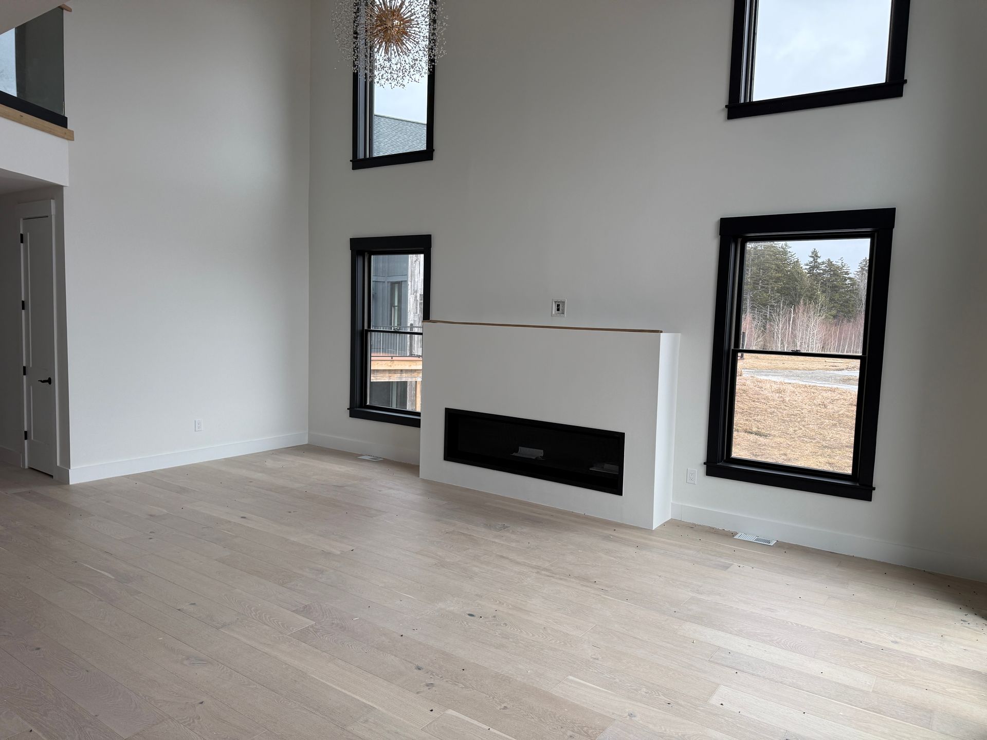A modern room with high ceilings, light wood floors, a white fireplace, and black-framed windows against white walls.