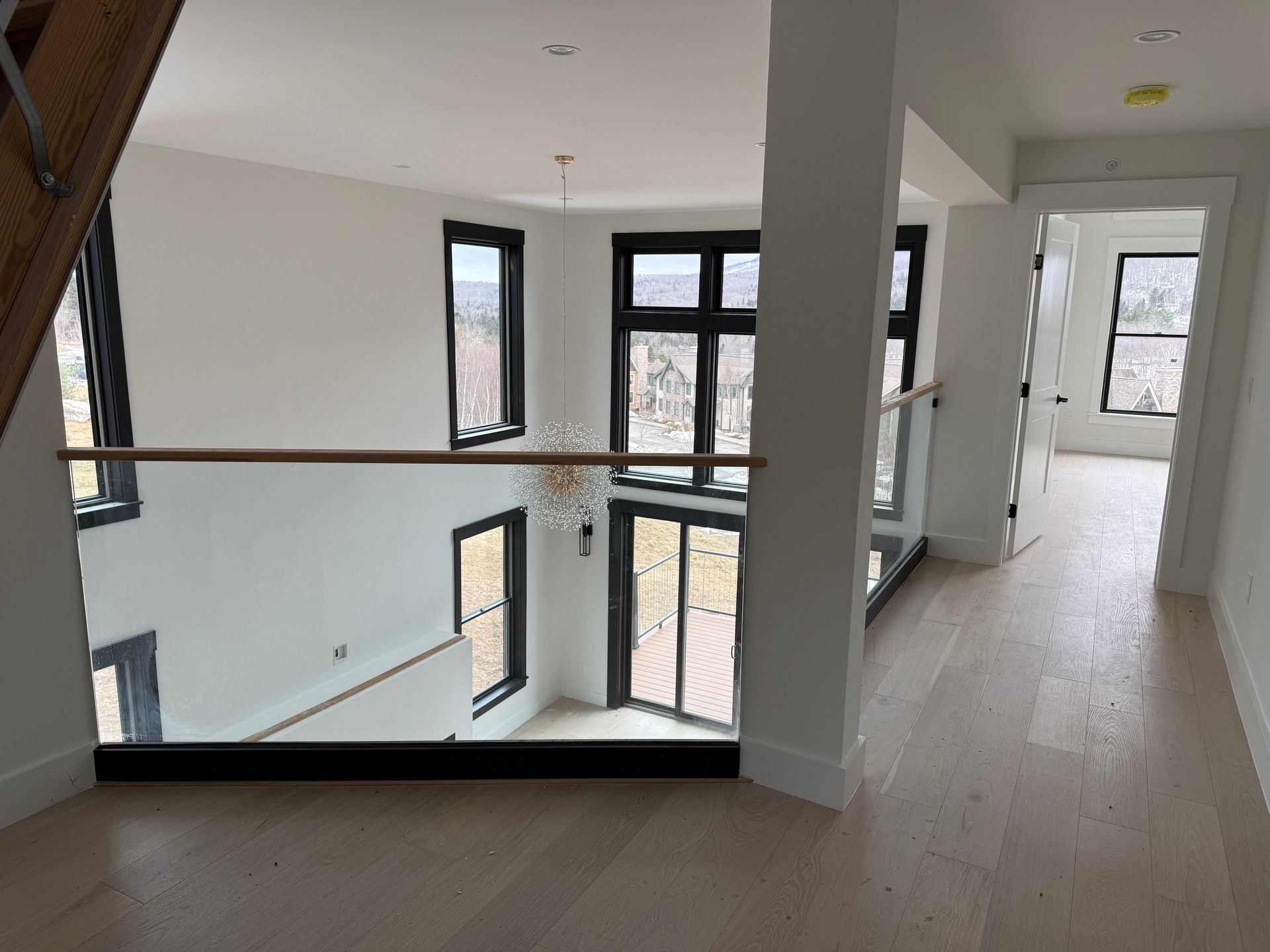 A bright, modern interior hallway overlooking a two-story living area with large black-framed windows and wood flooring.