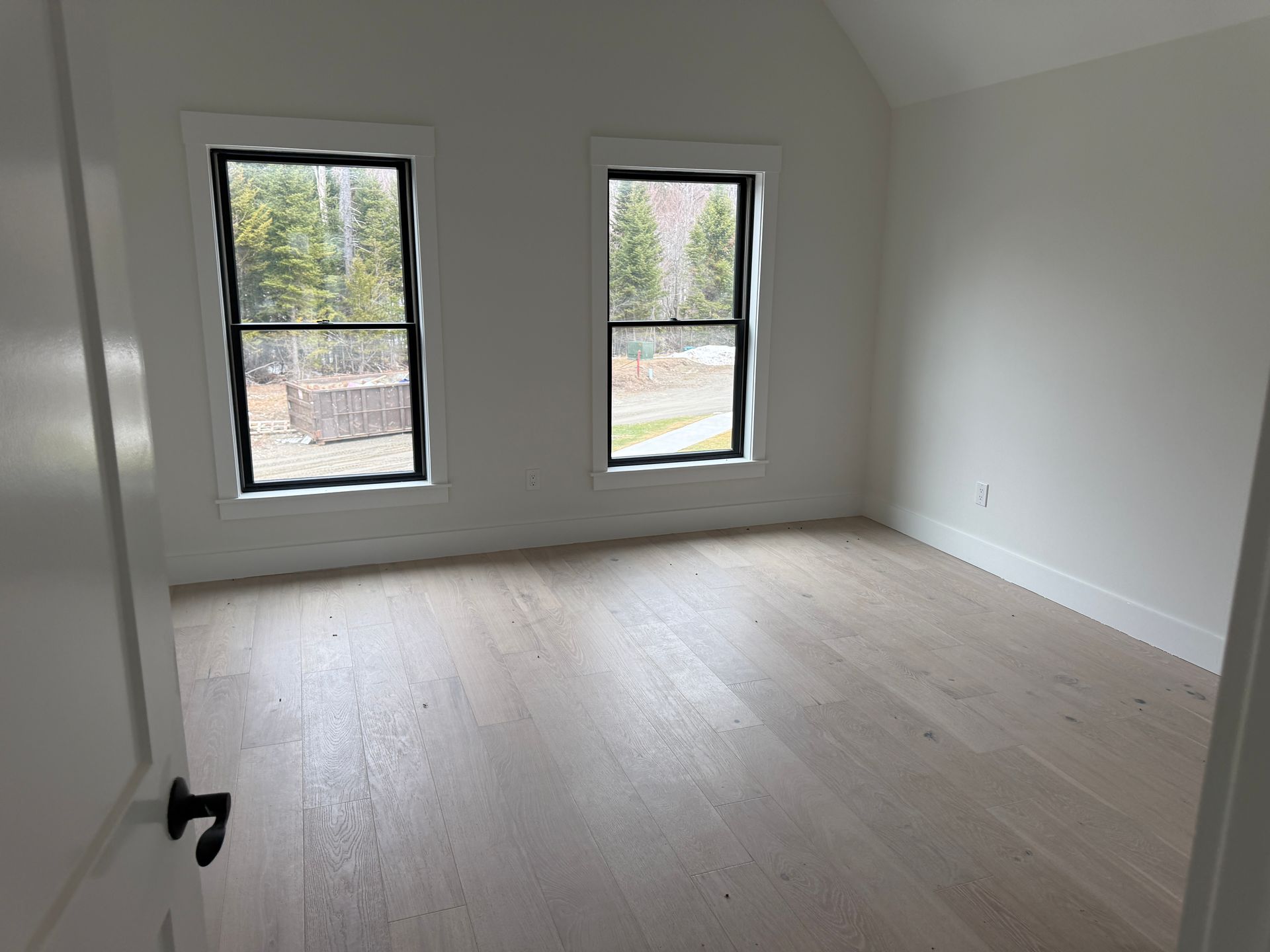 An empty room with light wood floors, white walls, and two windows looking out onto a wooded area.