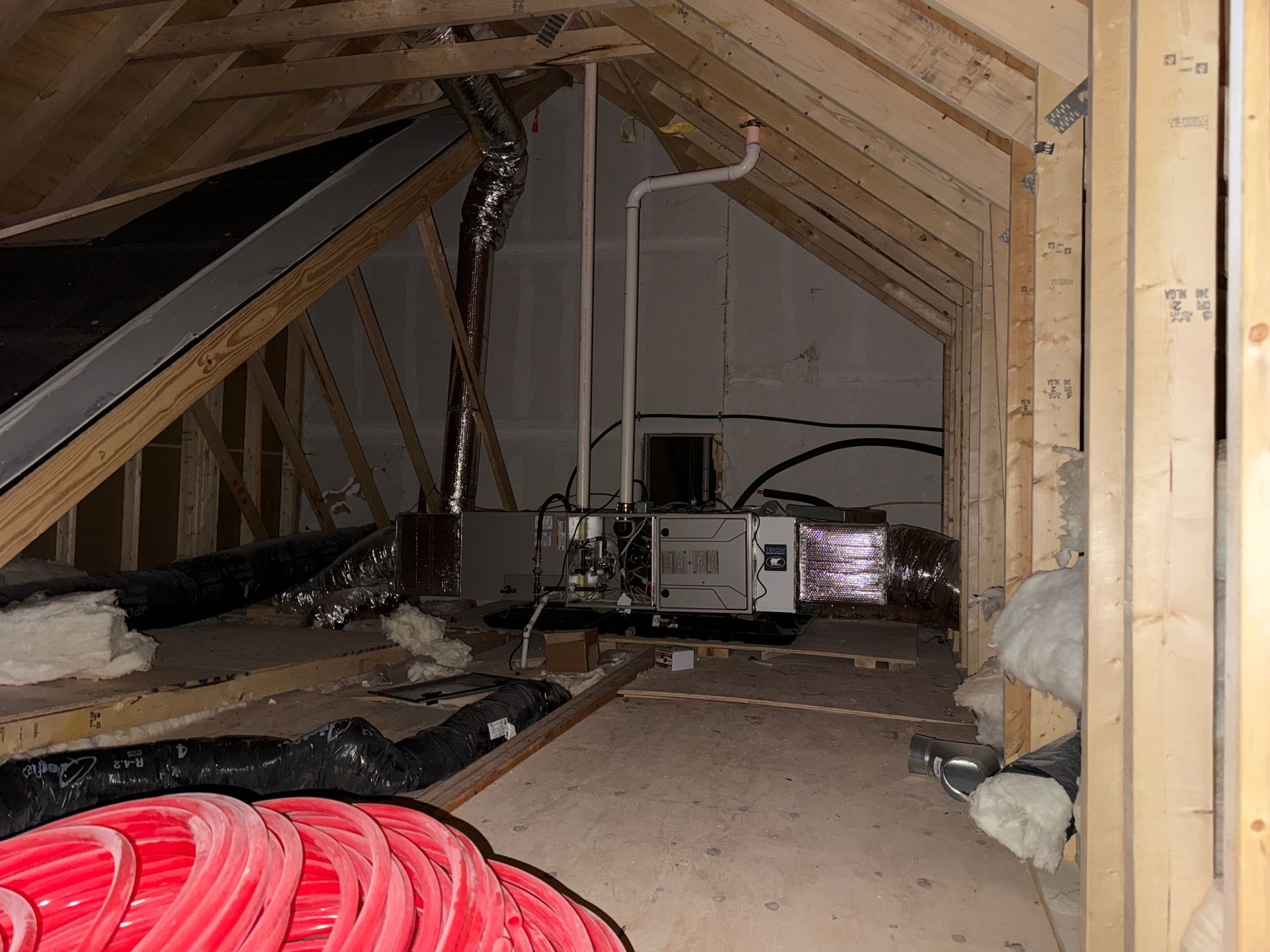 An HVAC air handler unit sits in an attic with exposed wooden rafters, white pipes, and red PEX tubing.