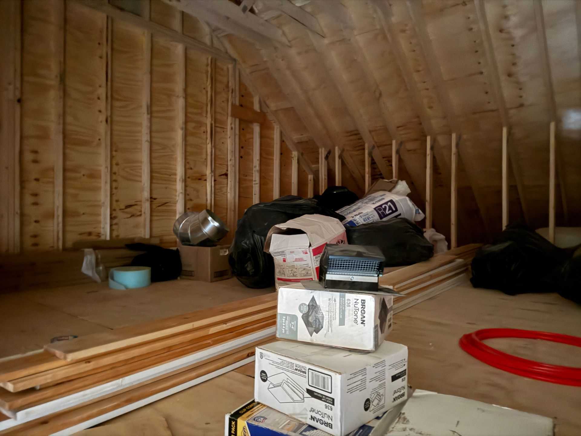An attic space with plywood walls and flooring, featuring stacked cardboard boxes, construction materials, and bags.