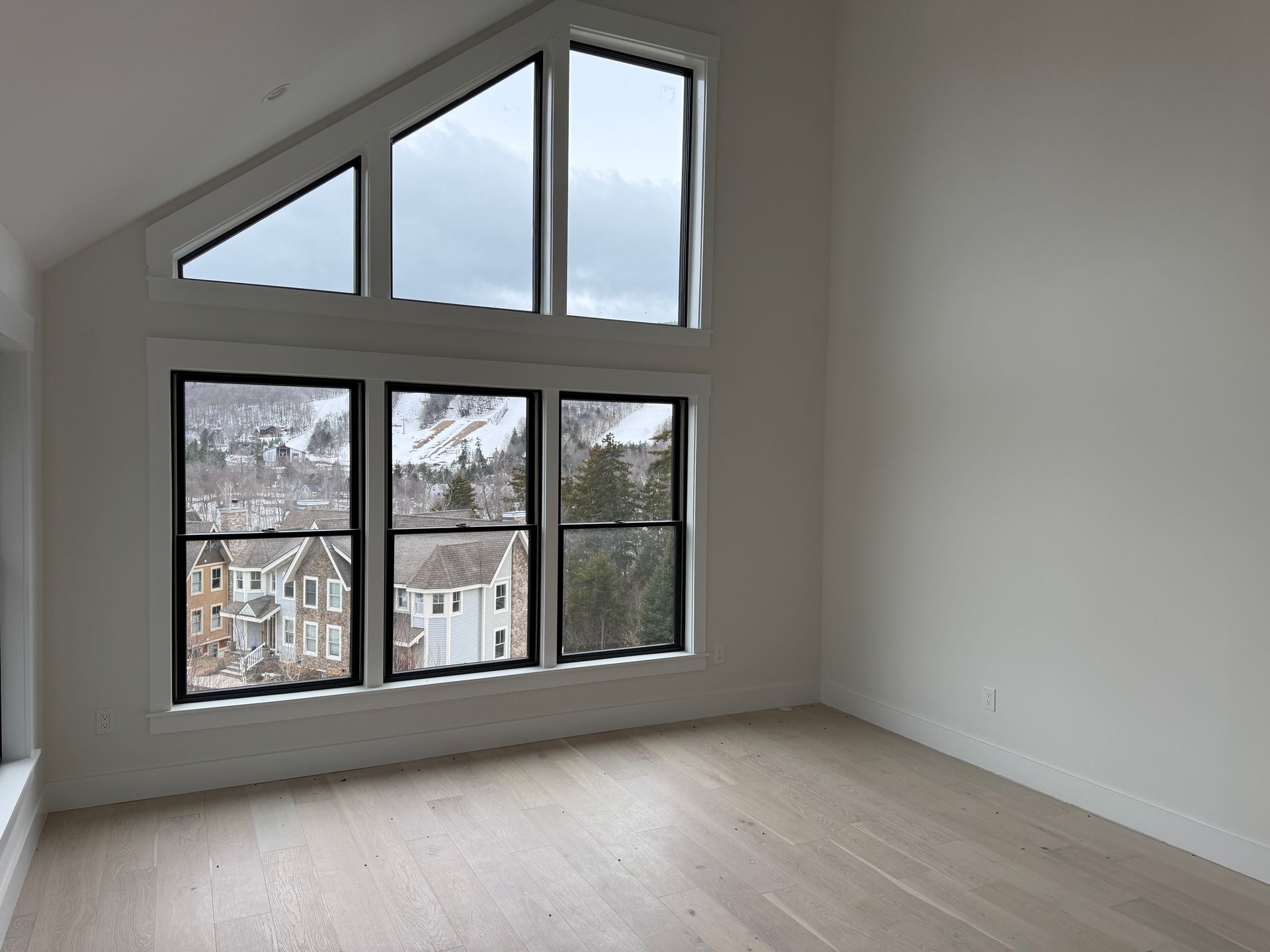 An empty, modern room with light wood floors, white walls, and a large window wall looking out at a snowy residential area.