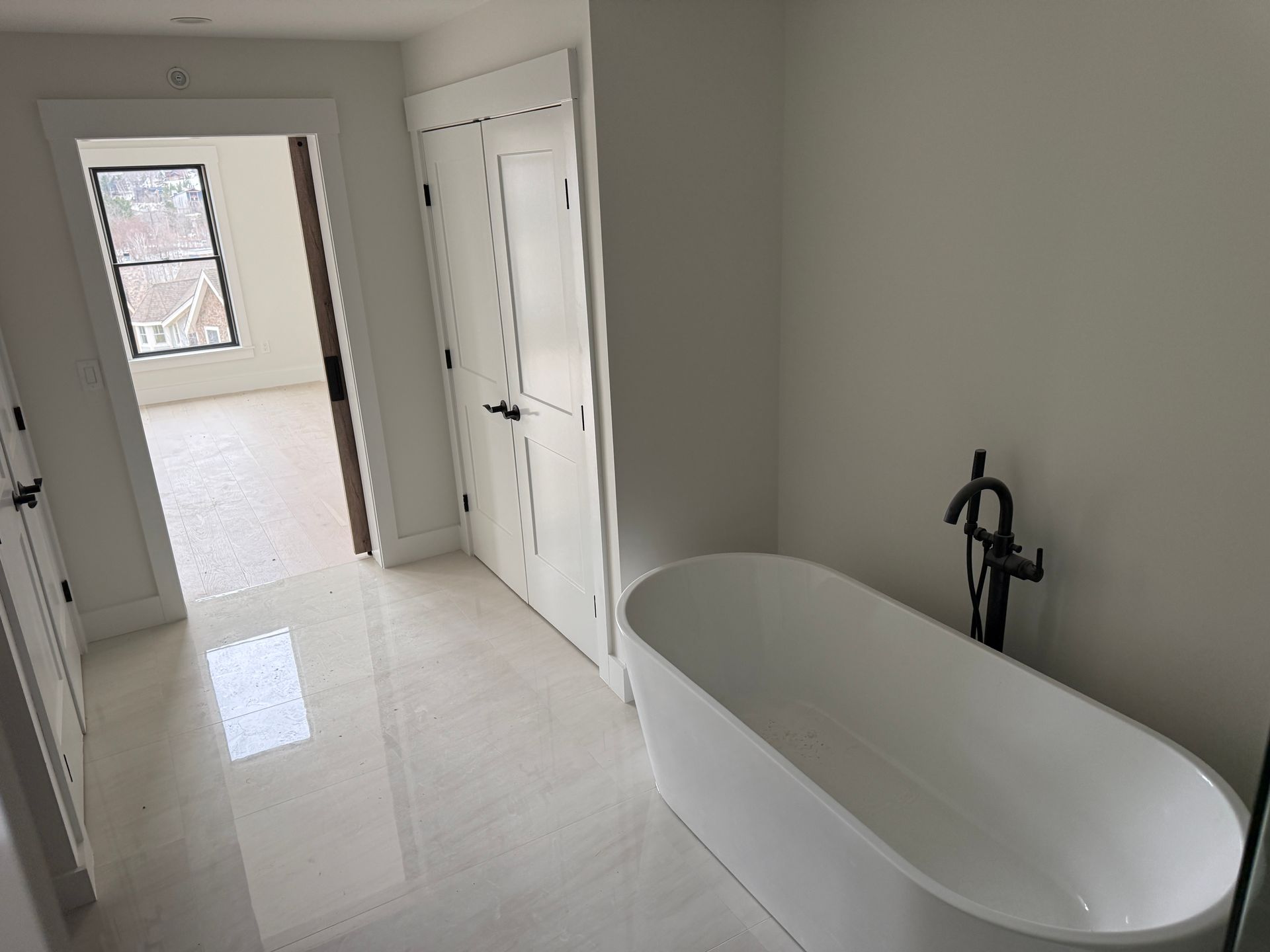 A white freestanding bathtub with a black faucet in a modern bathroom with light tile floors and white double doors.