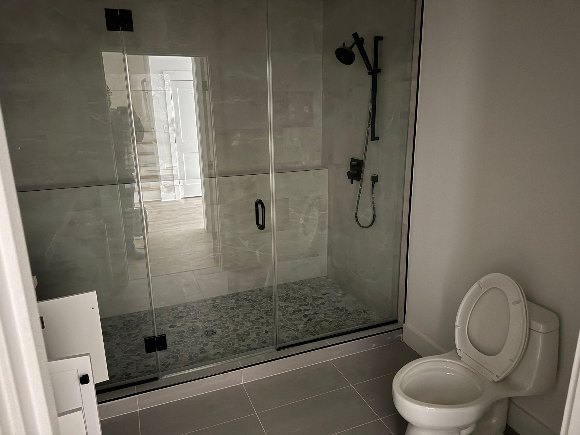 A bathroom featuring a glass-enclosed shower with pebbled floor tiles next to a white toilet on light grey floor tiles.