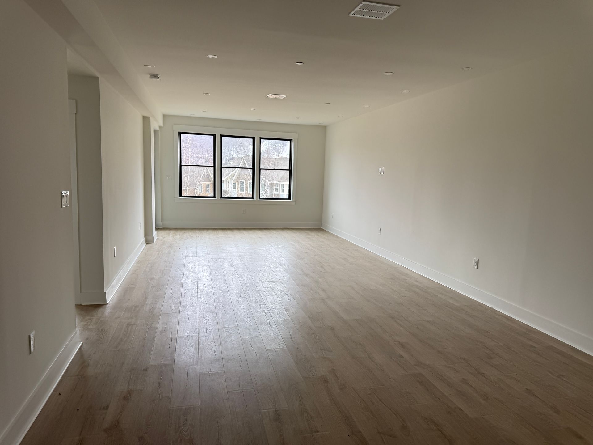 An empty, rectangular room with light wood-tone flooring, white walls, and a large triple window at the far end.