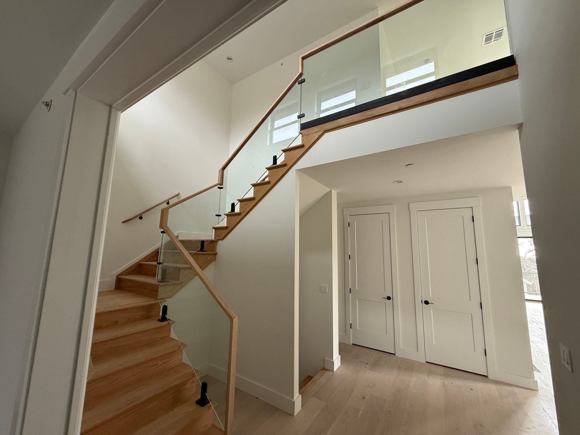 A wooden staircase with a glass railing ascends to an upper level next to two white closed doors in a modern home.