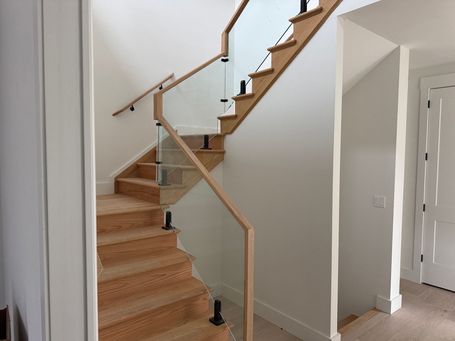 Modern wooden staircase with glass railings and black supports in a white-walled interior.