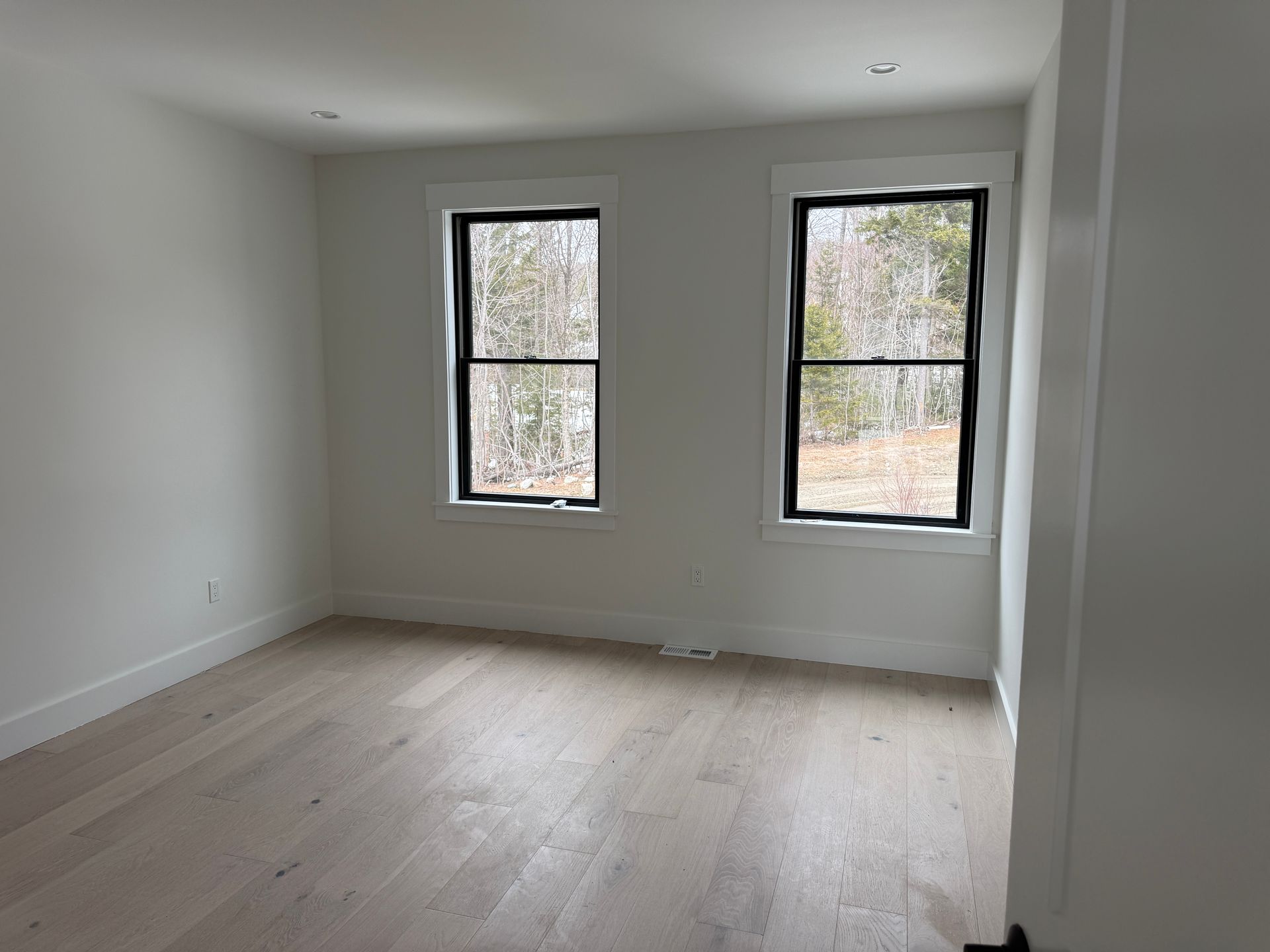 An empty room with light wood floors, neutral walls, two windows looking onto trees, and recessed ceiling lights.