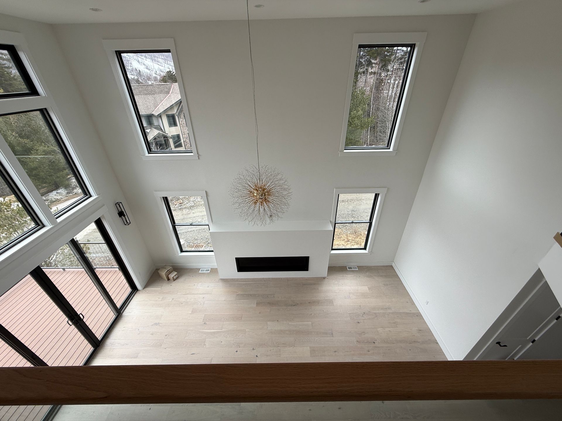High-ceilinged living room with light wood floors, a central fireplace, a modern chandelier, and large window panels.