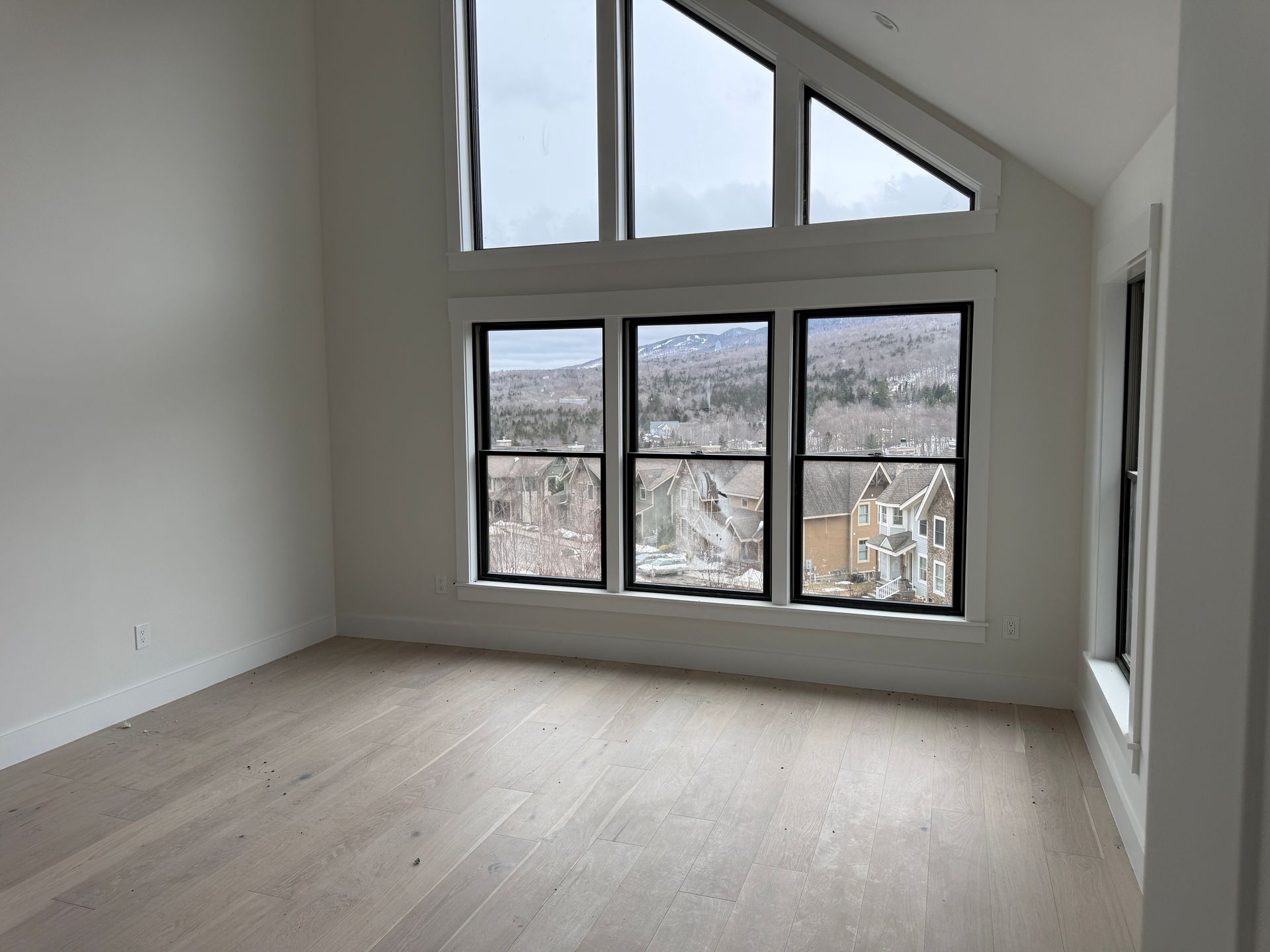A room with light wood floors, white walls, and a large window arrangement overlooking a town and mountains.