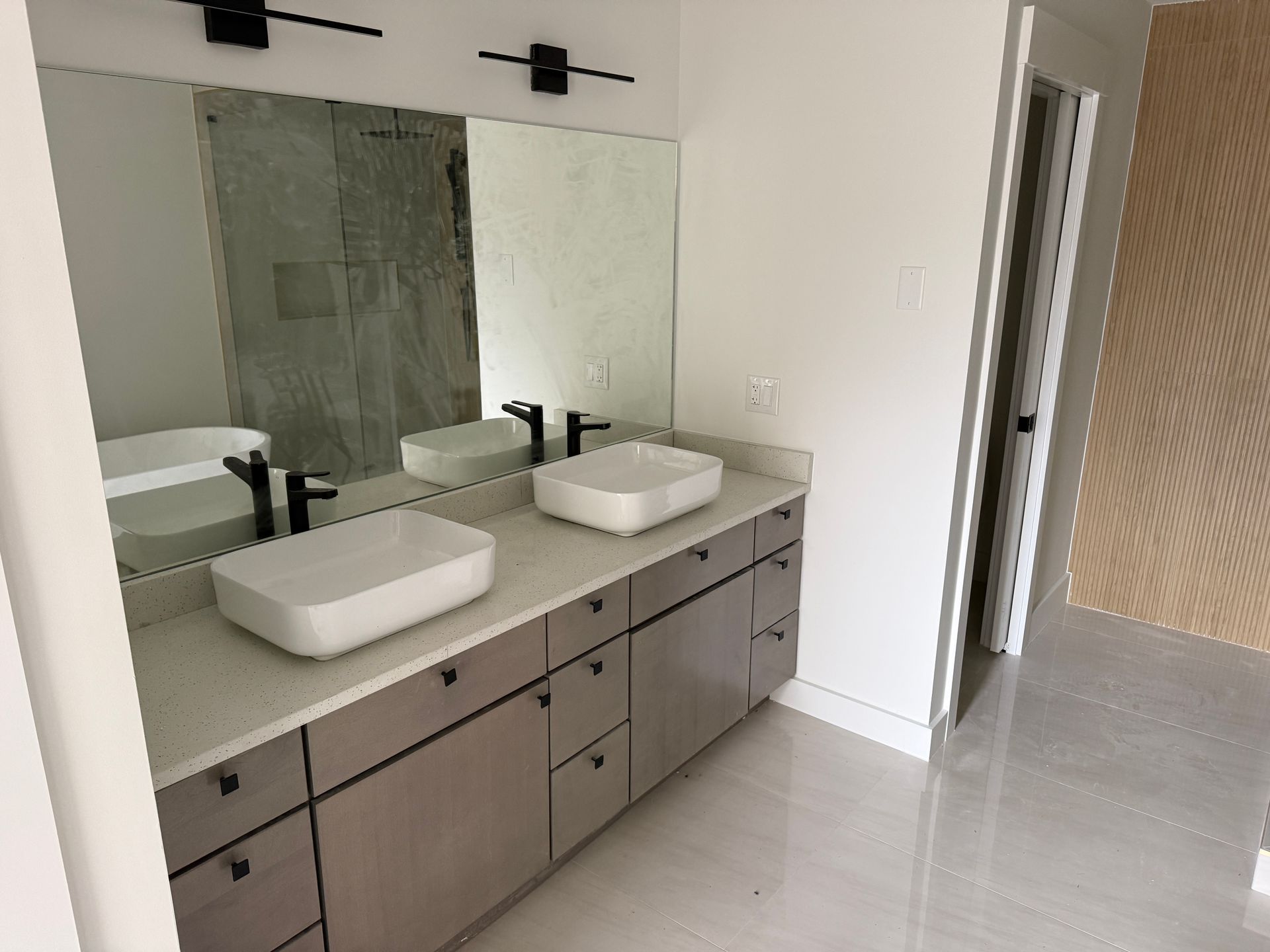 Modern bathroom vanity with two white vessel sinks, matte black faucets, grey cabinets, and a large wall-to-wall mirror.