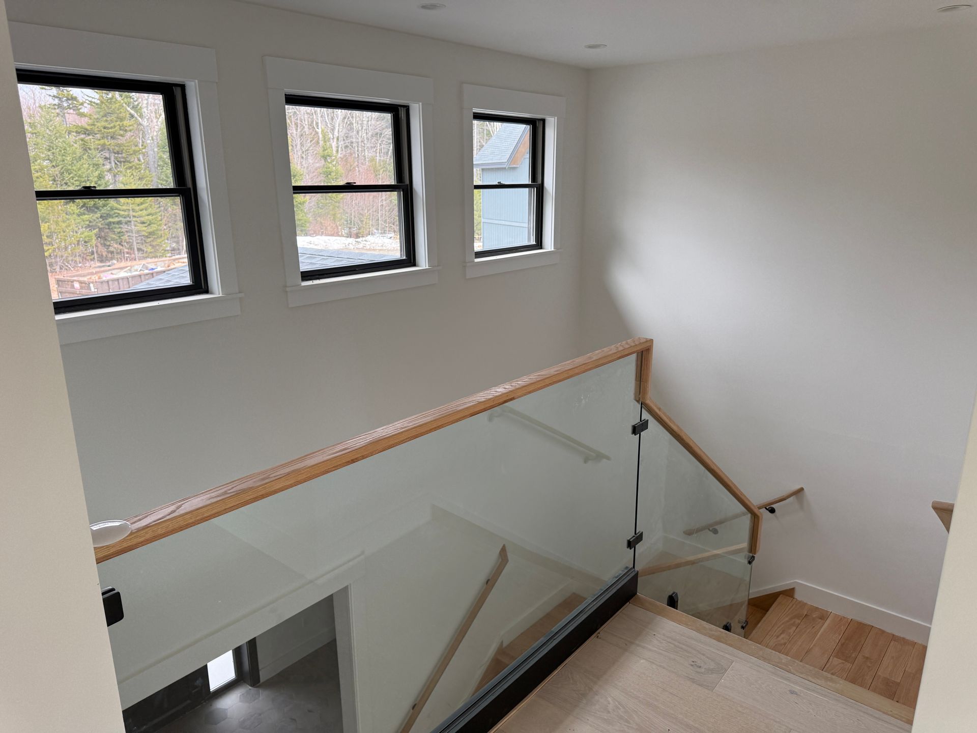 A modern staircase with a glass railing and wooden handrail, leading down near a wall with three white-framed windows.