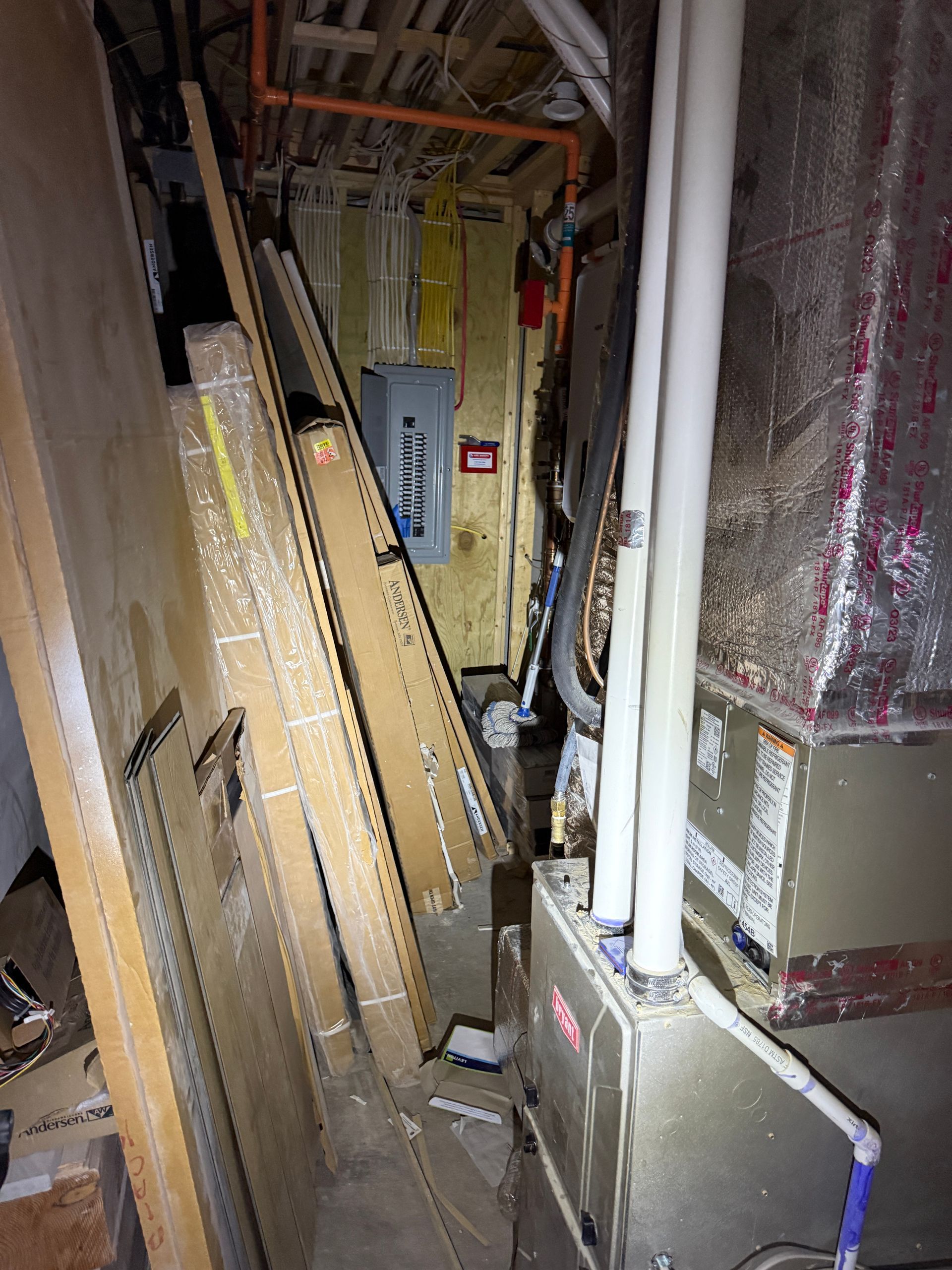 A narrow basement utility area featuring a furnace, exposed white pipes, a grey electrical panel, and stacked lumber.