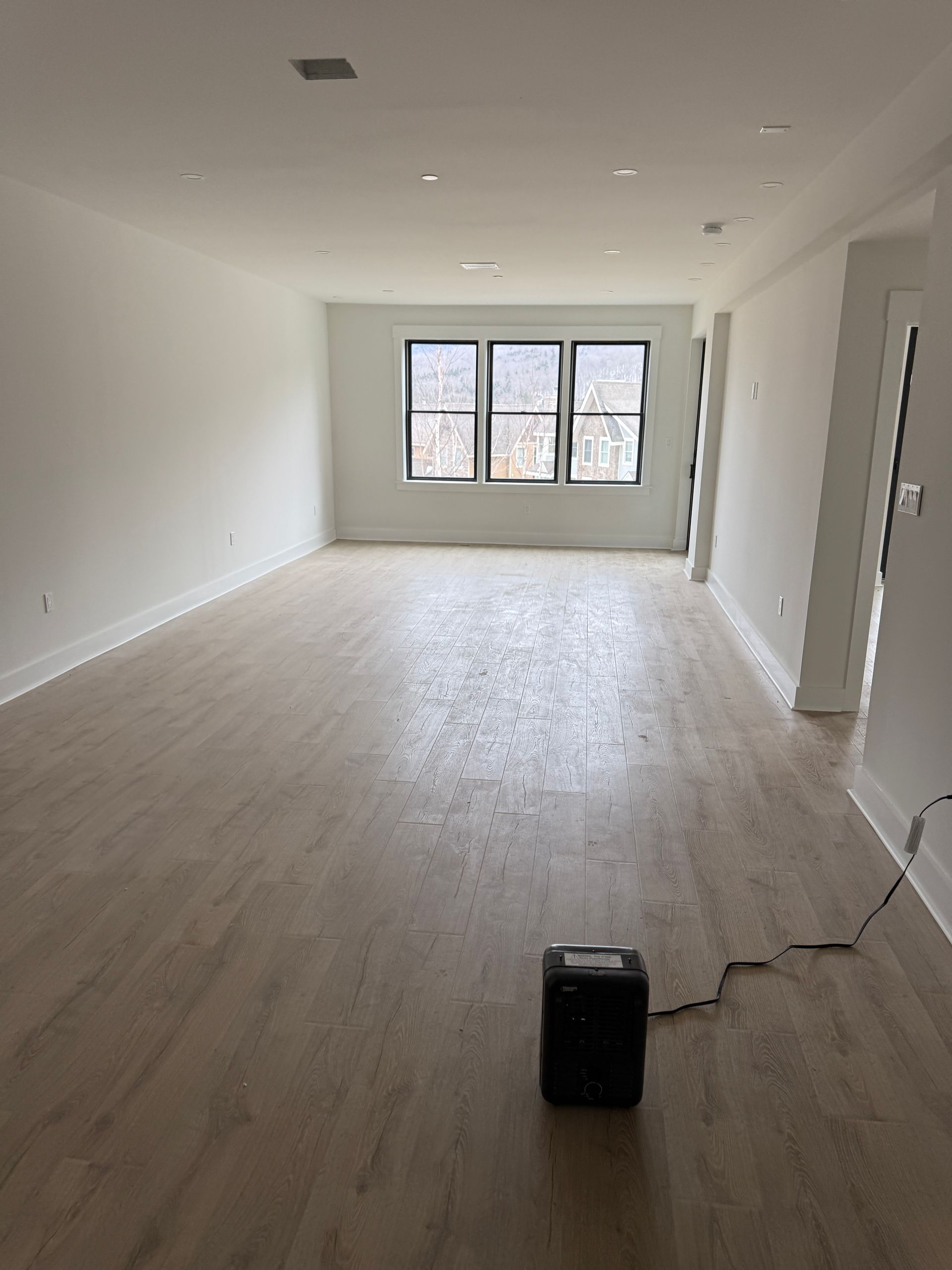 A bright, empty room with light wood-look flooring, white walls, and a large window, with a small black heater on the floor.