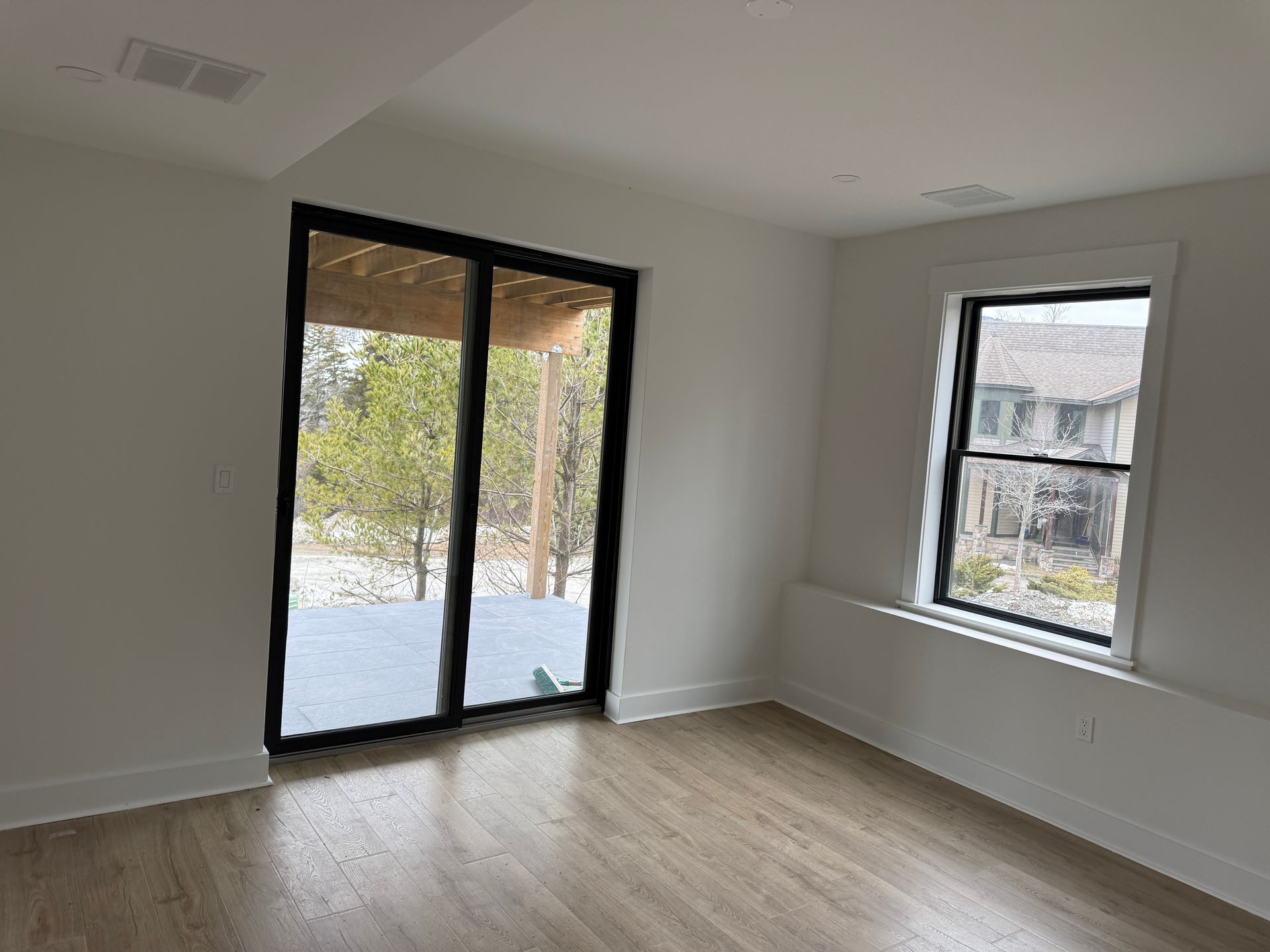 An empty room with white walls, light wood floors, a sliding glass door to a deck, and a window looking out at trees.