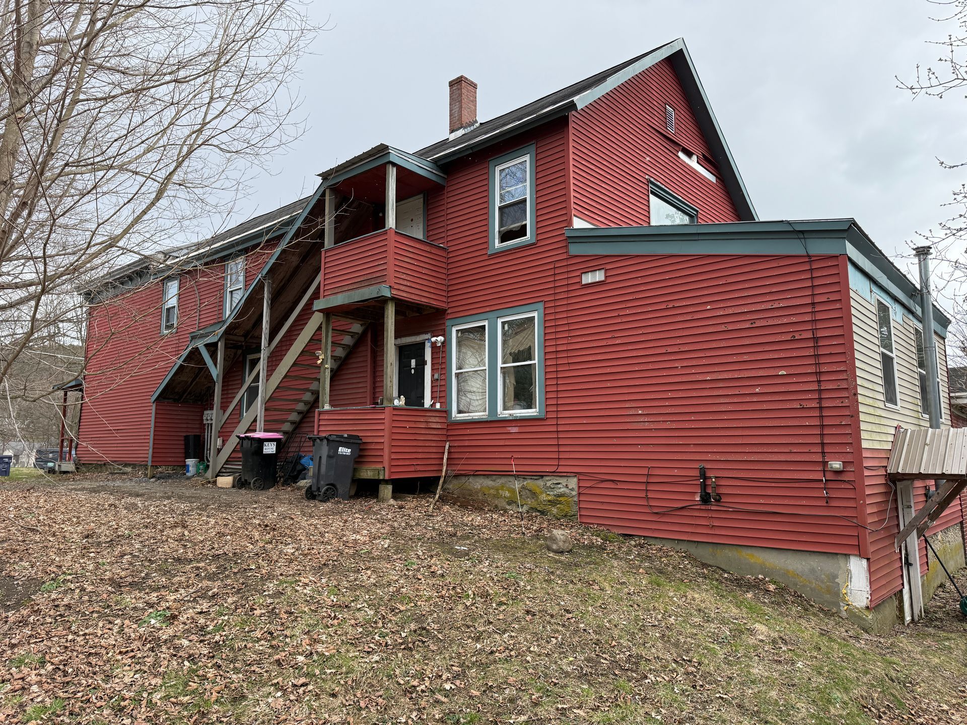 A red two-story house on a slight hill featuring a wooden exterior staircase, a chimney, and a small porch.