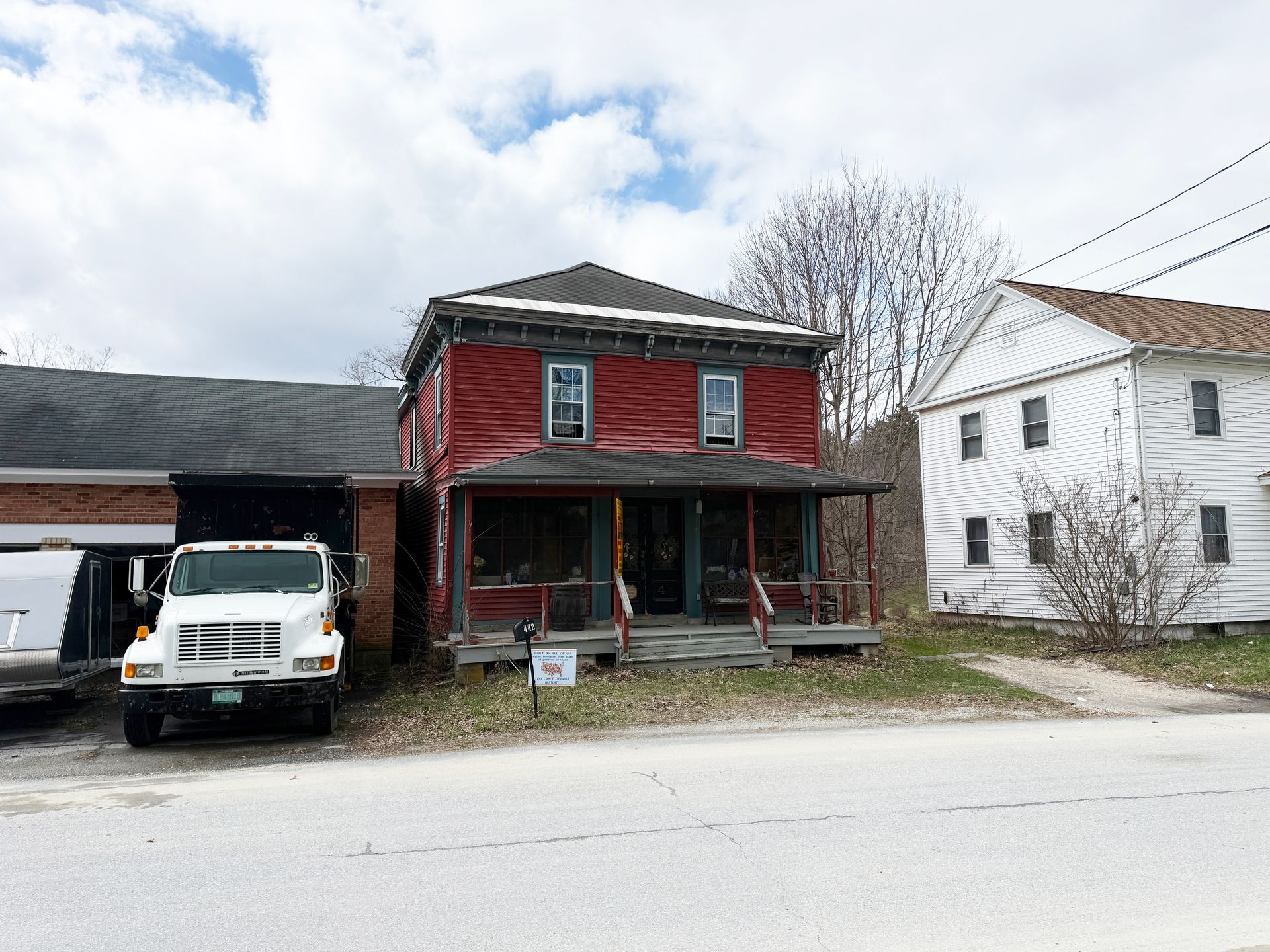 A red two-story house with a front porch sits beside a white building and a parked white truck on a paved road.