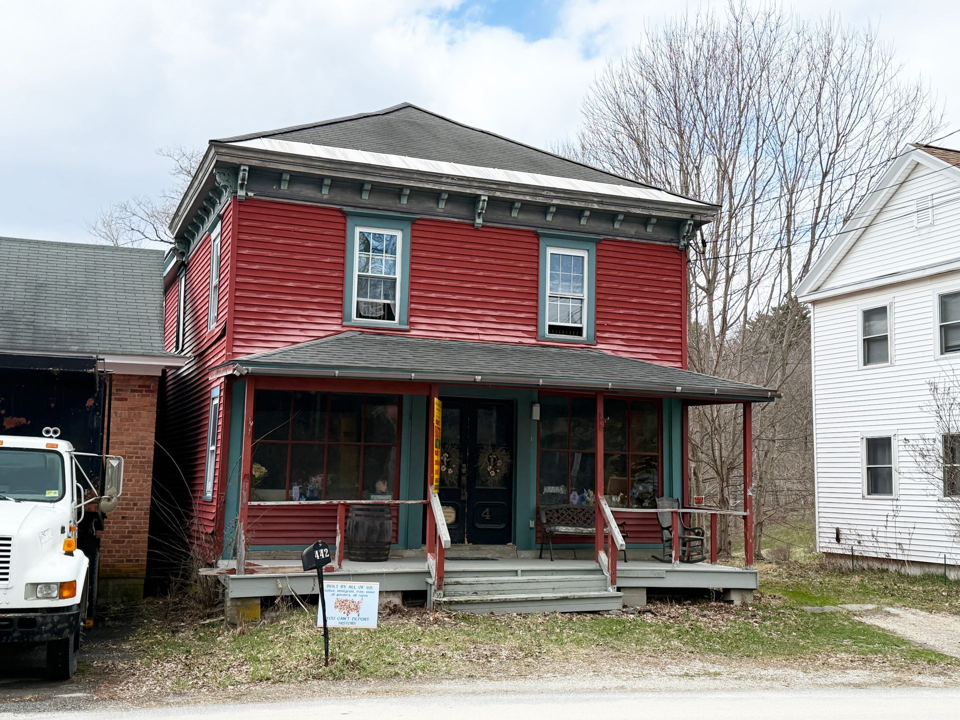 A two-story red building with a porch and dark trim stands next to a white house, with a white truck parked to the left.