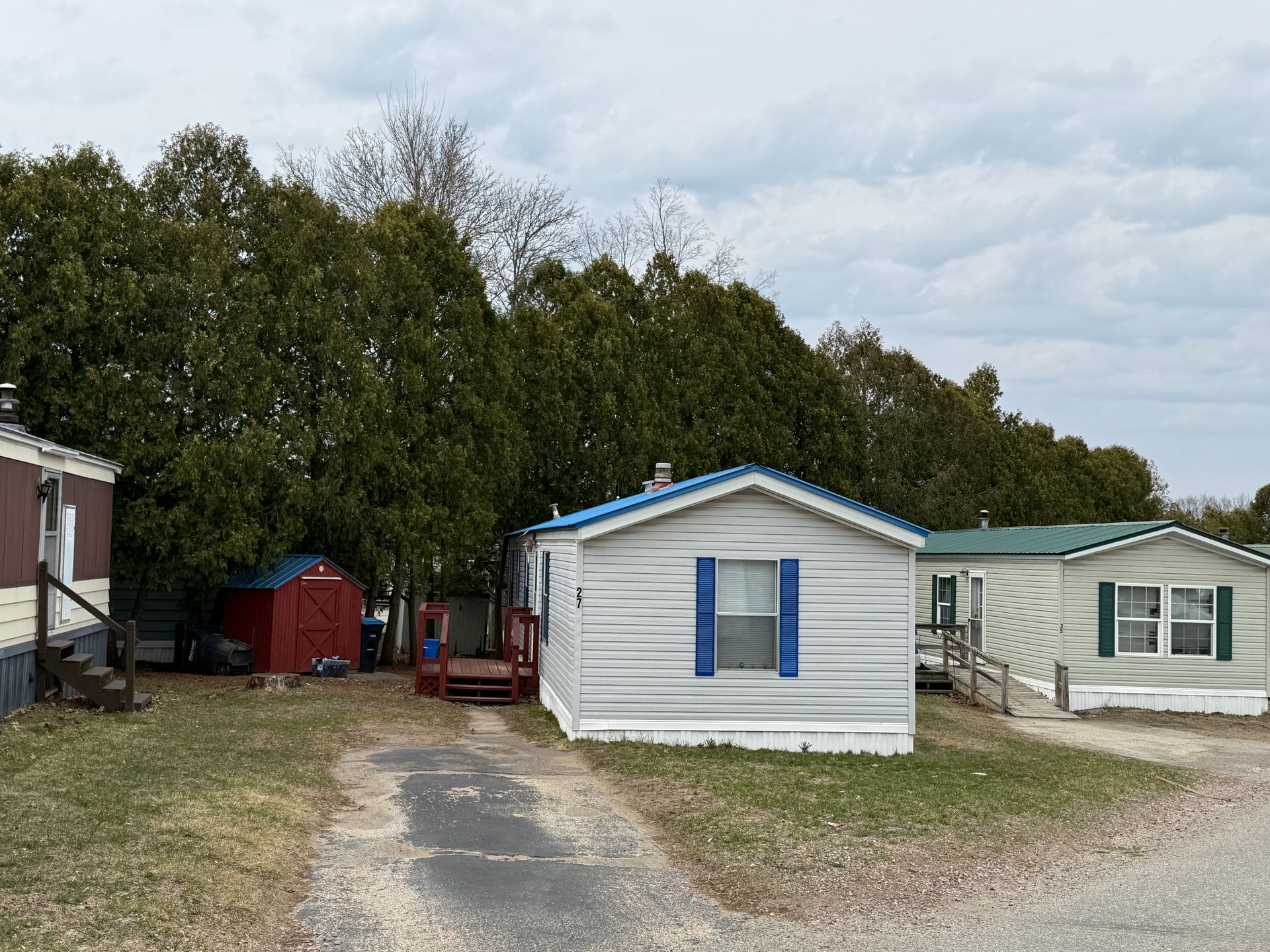 Several manufactured homes with light-colored siding, blue shutters, and a small red shed in a grassy, tree-lined area.