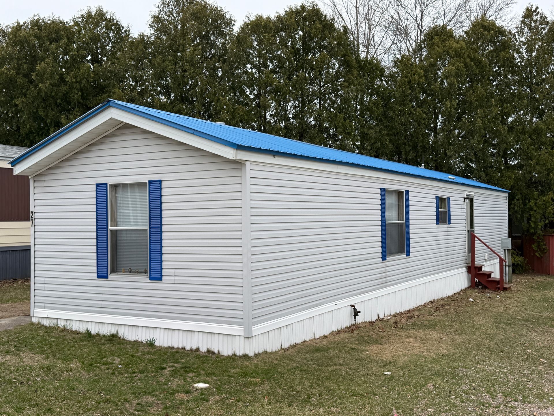 A light gray mobile home with a bright blue metal roof, blue shutters, and stairs leading to a side door.