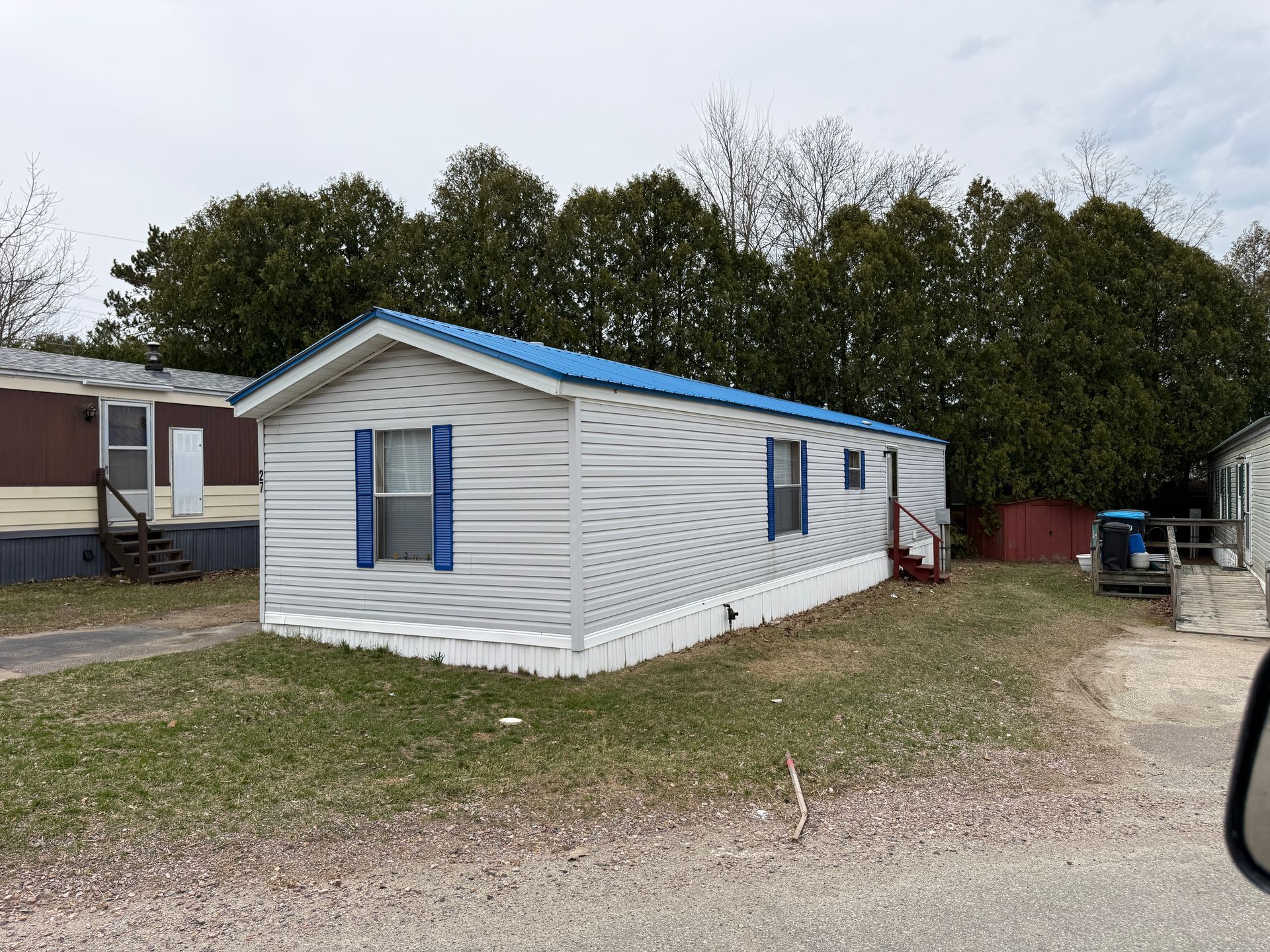 A white mobile home with a blue roof and blue shutters stands on a grassy lot in front of a row of tall trees.