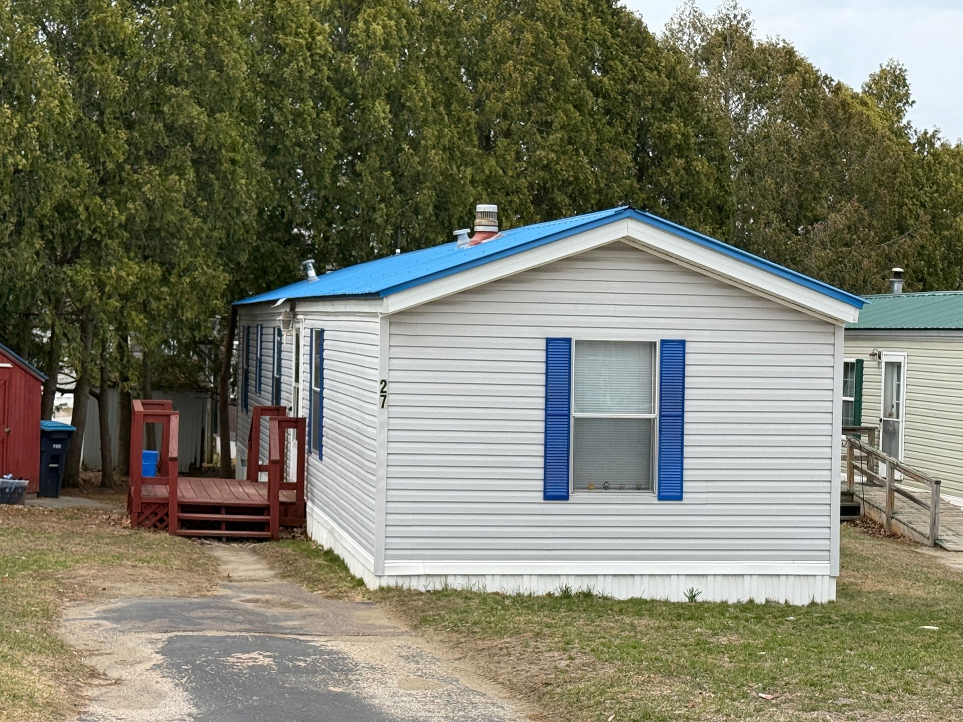 A single-story mobile home with light gray siding, a bright blue metal roof, and blue shutters, sitting on a gravel lot.