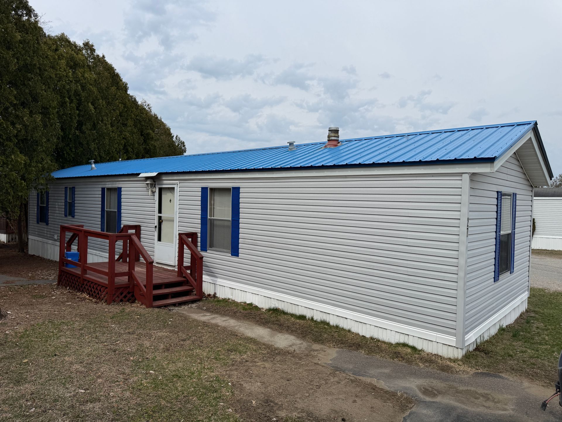 A single-story mobile home with gray horizontal siding, blue shutters, and a bright blue metal roof, featuring a small deck.