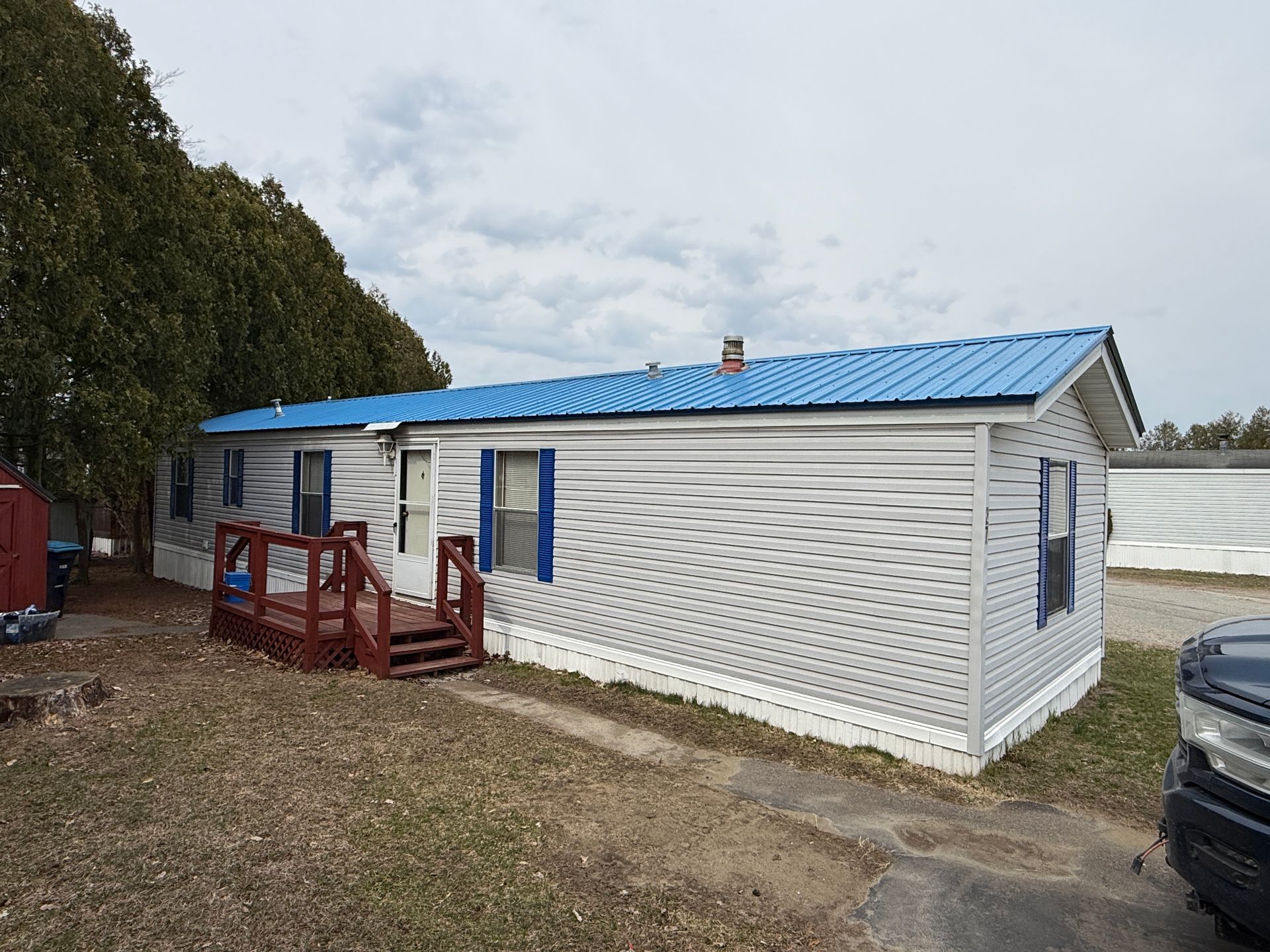 A single-story manufactured home with gray horizontal siding, a bright blue metal roof, and a small red wooden deck.
