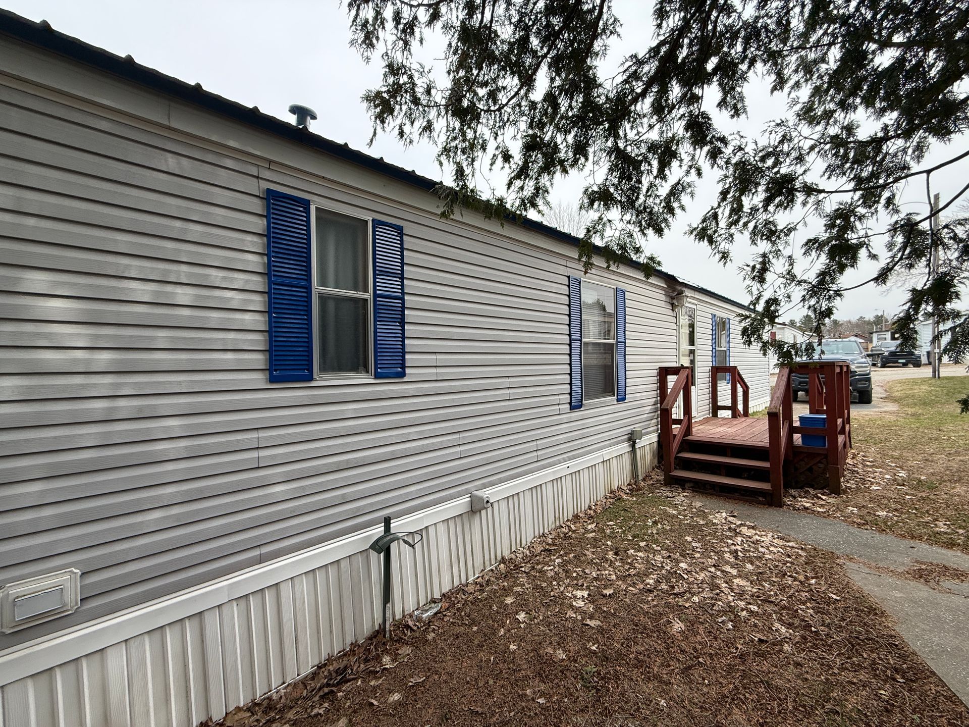 A single-story, light-colored mobile home with blue shutters, a brown wooden deck, and white skirting on a gravel lot.