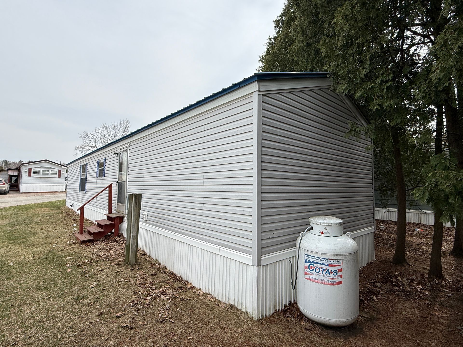 A mobile home with gray horizontal siding and white skirting, featuring a wooden staircase and a white propane tank nearby.