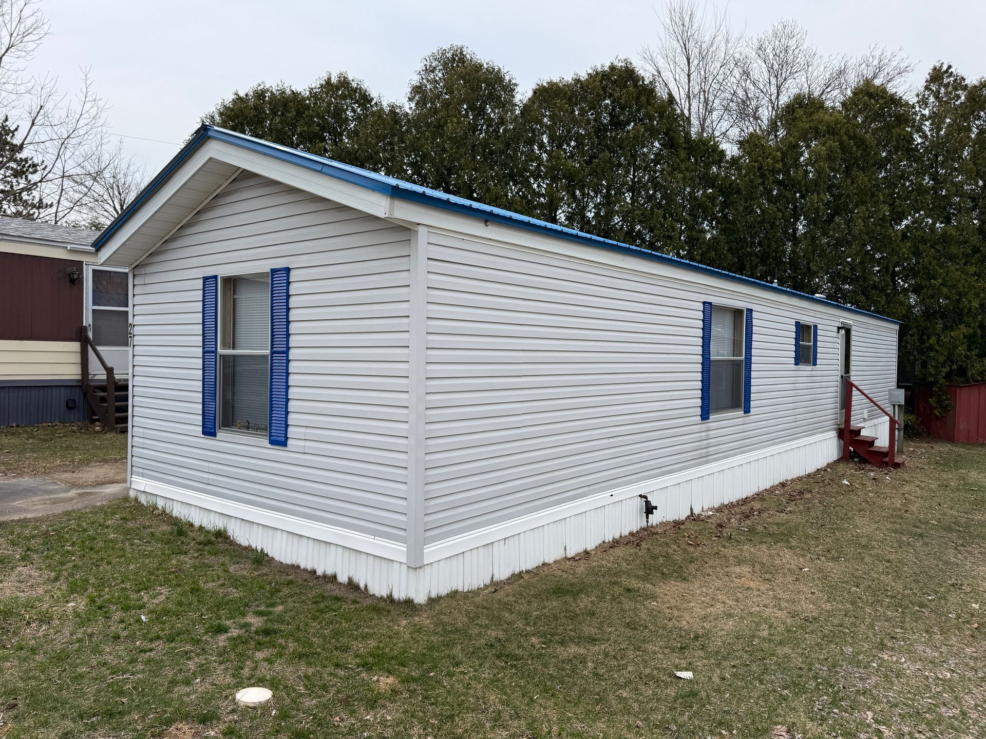 A light-gray mobile home with a blue roof, blue window shutters, and white skirting sits on a lawn before evergreen trees.