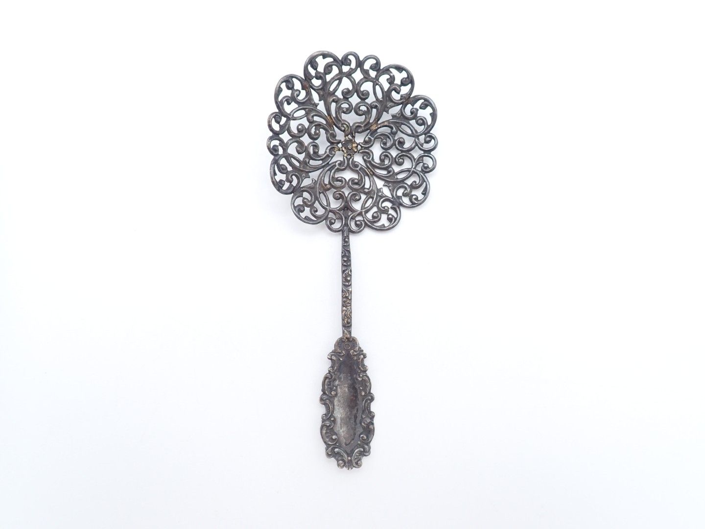 A decorative silver utensil with an ornate circular top and dangling teardrop design on a white background