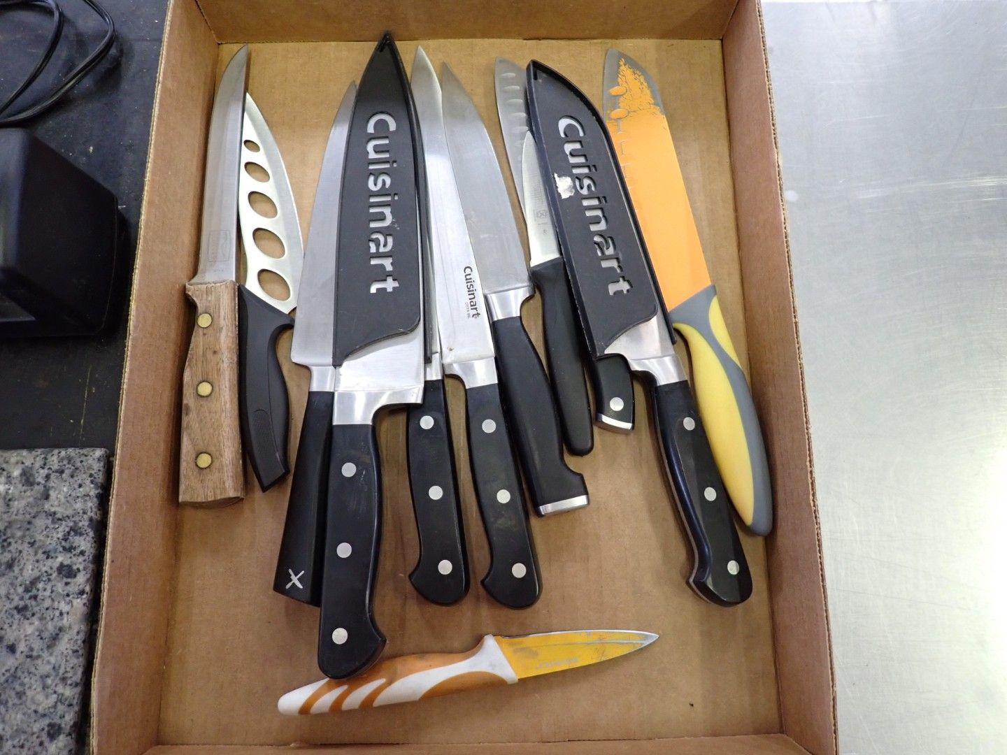 Kitchen knives arranged in a cardboard box, including several Cuisinart knives and a peeler.
