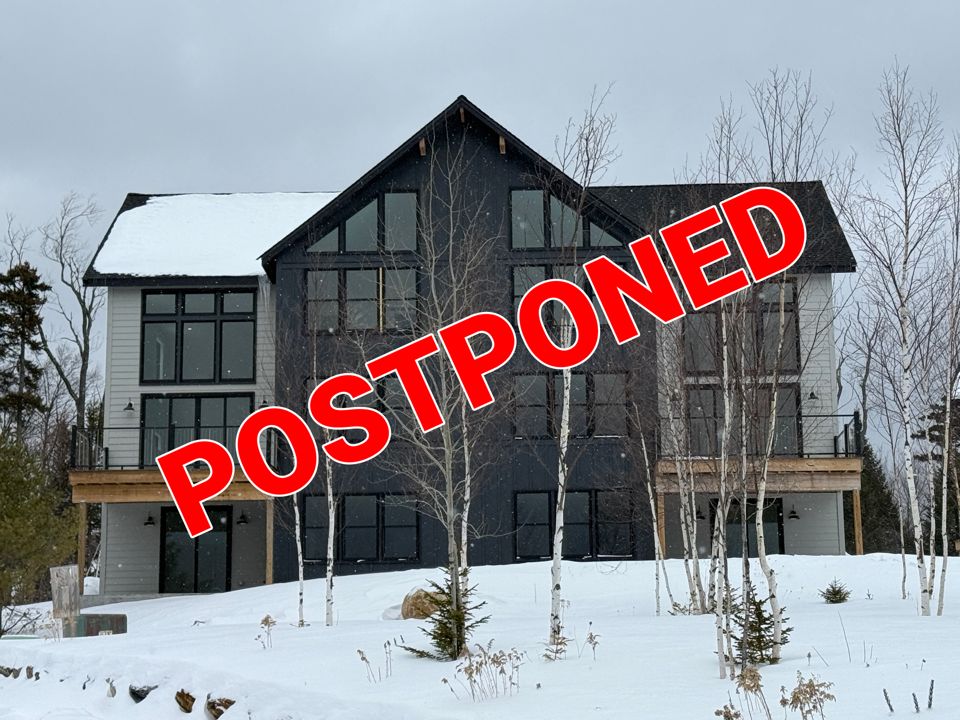 A three-story, modern house with grey and dark siding stands in a snowy, wooded landscape with birch trees.
