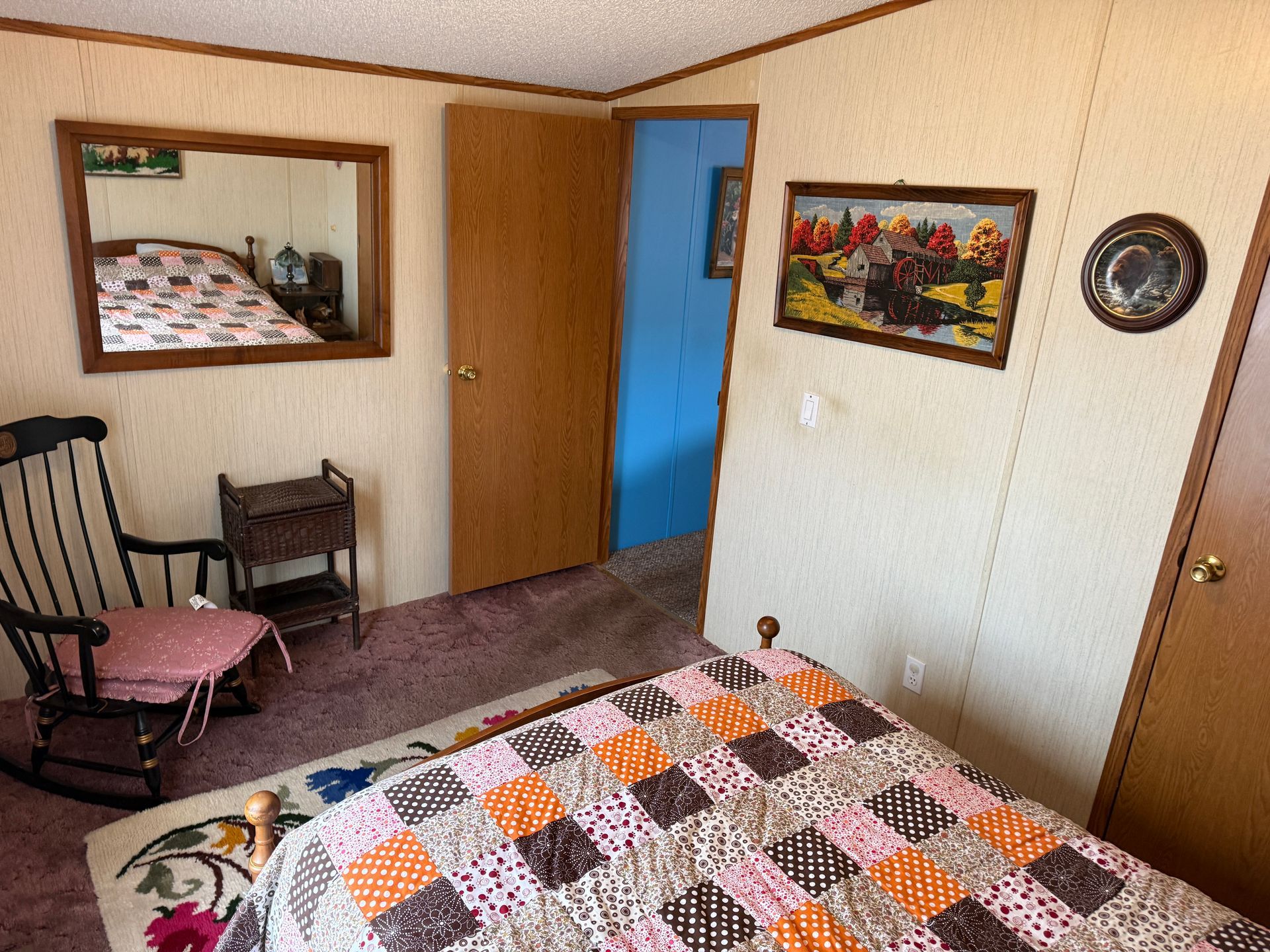 Small bedroom with a patterned bedspread, chair, mirror, open door, and wall art.