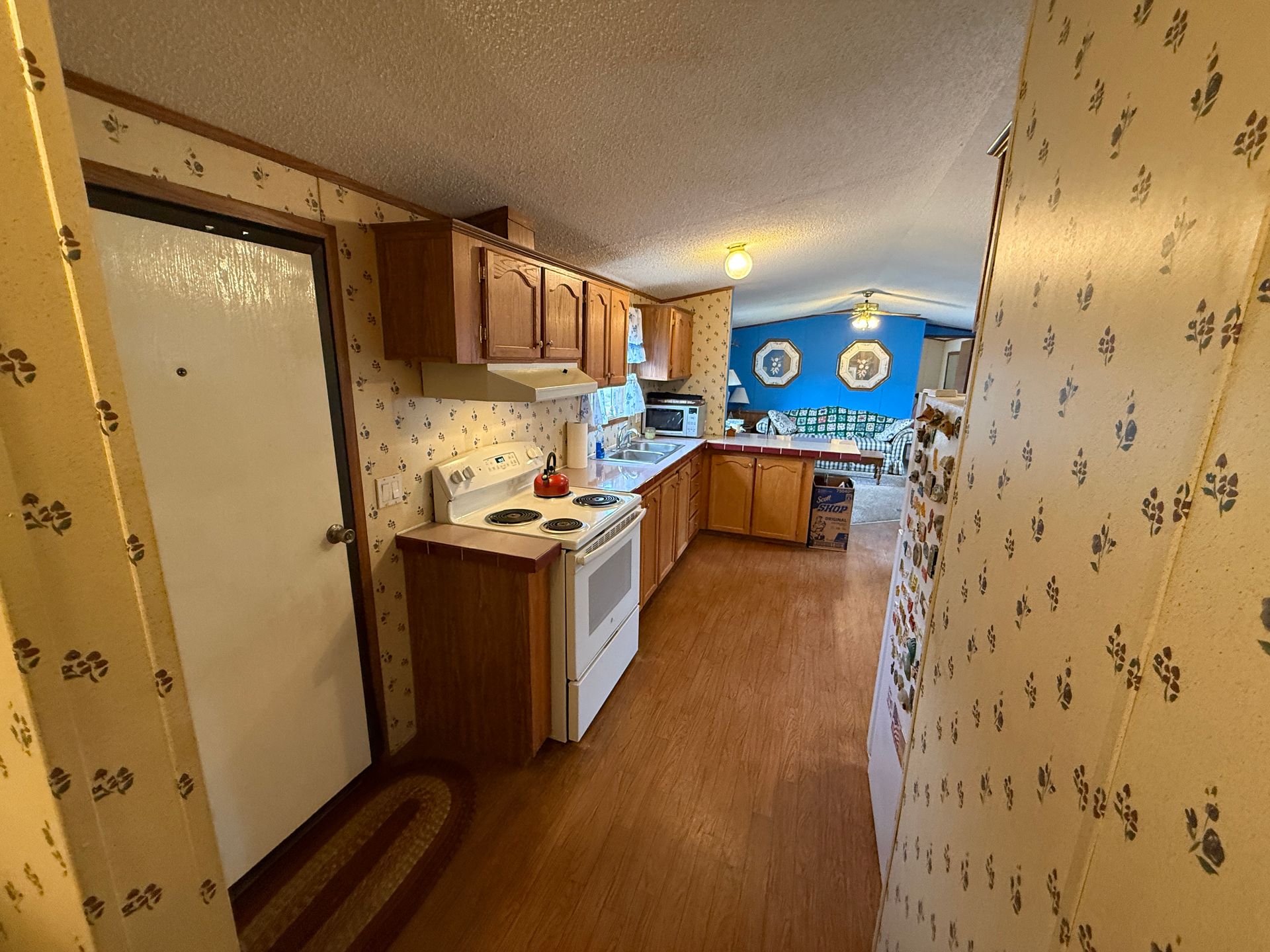 Narrow kitchen hallway with wood cabinets, white appliances, floral wallpaper, and a blue dining area ahead