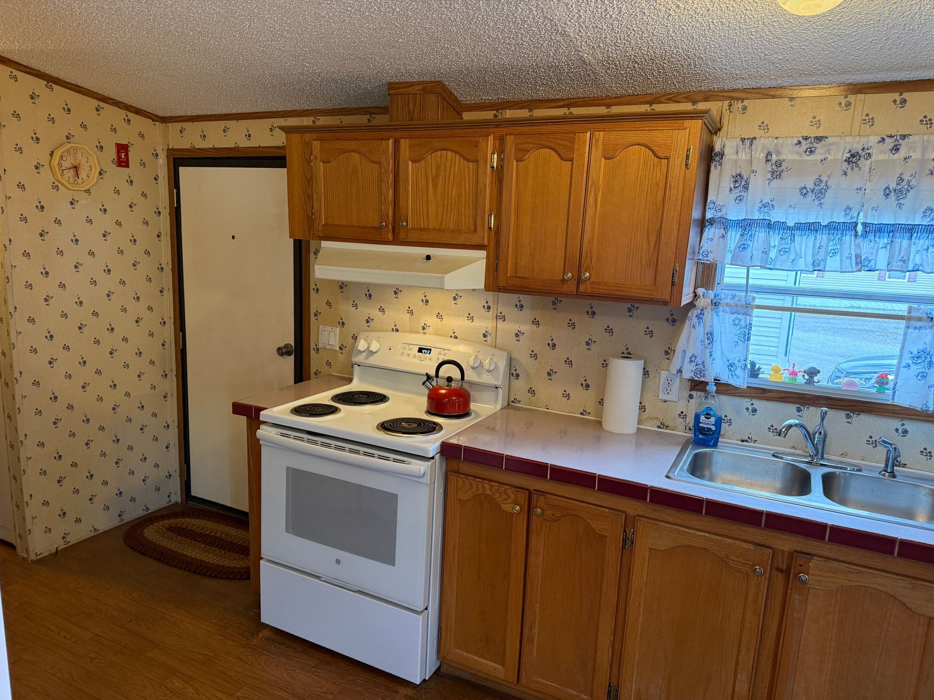 Small kitchen with white stove, wooden cabinets, sink, and blue curtains by the window.