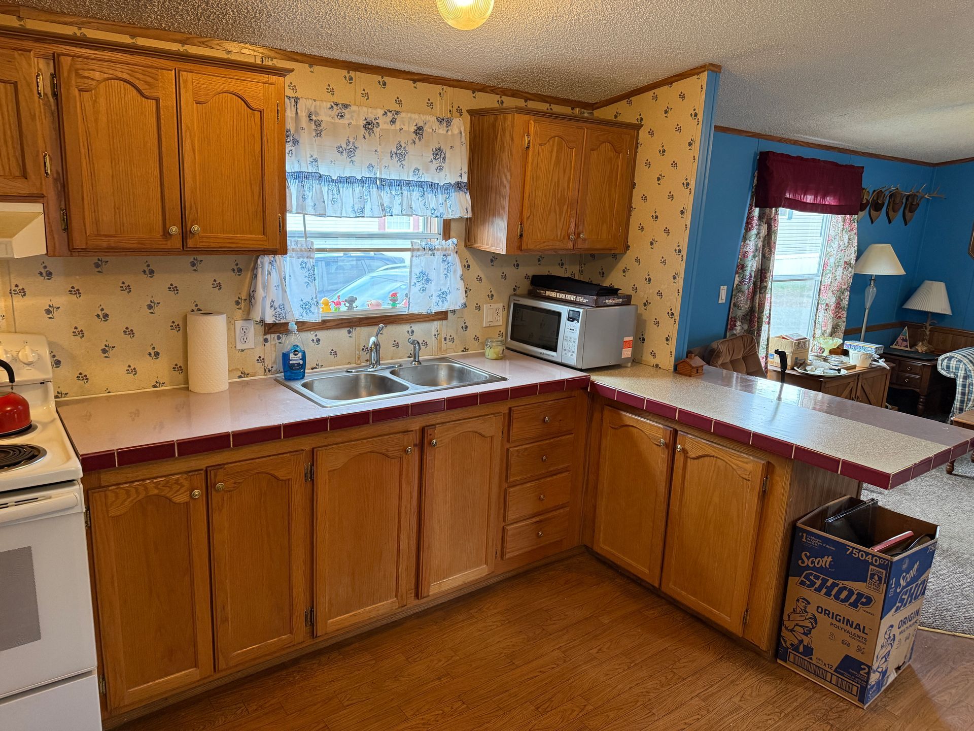 Small home kitchen with wood cabinets, beige counters, sink under window, and blue walls with a dining area beyond.