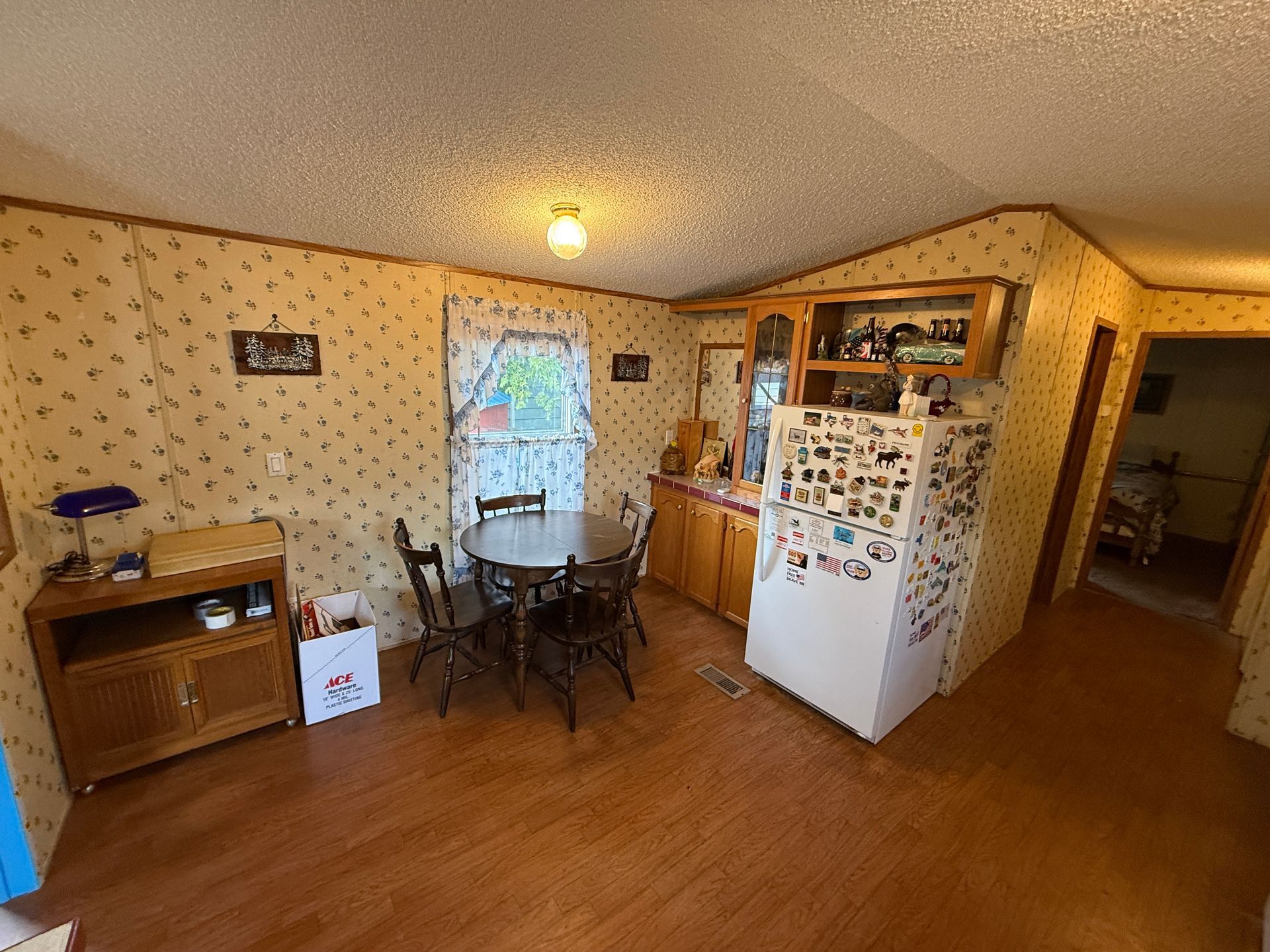 Small kitchen and dining area with wooden floor, white fridge, table, cabinets, and a doorway to another room.