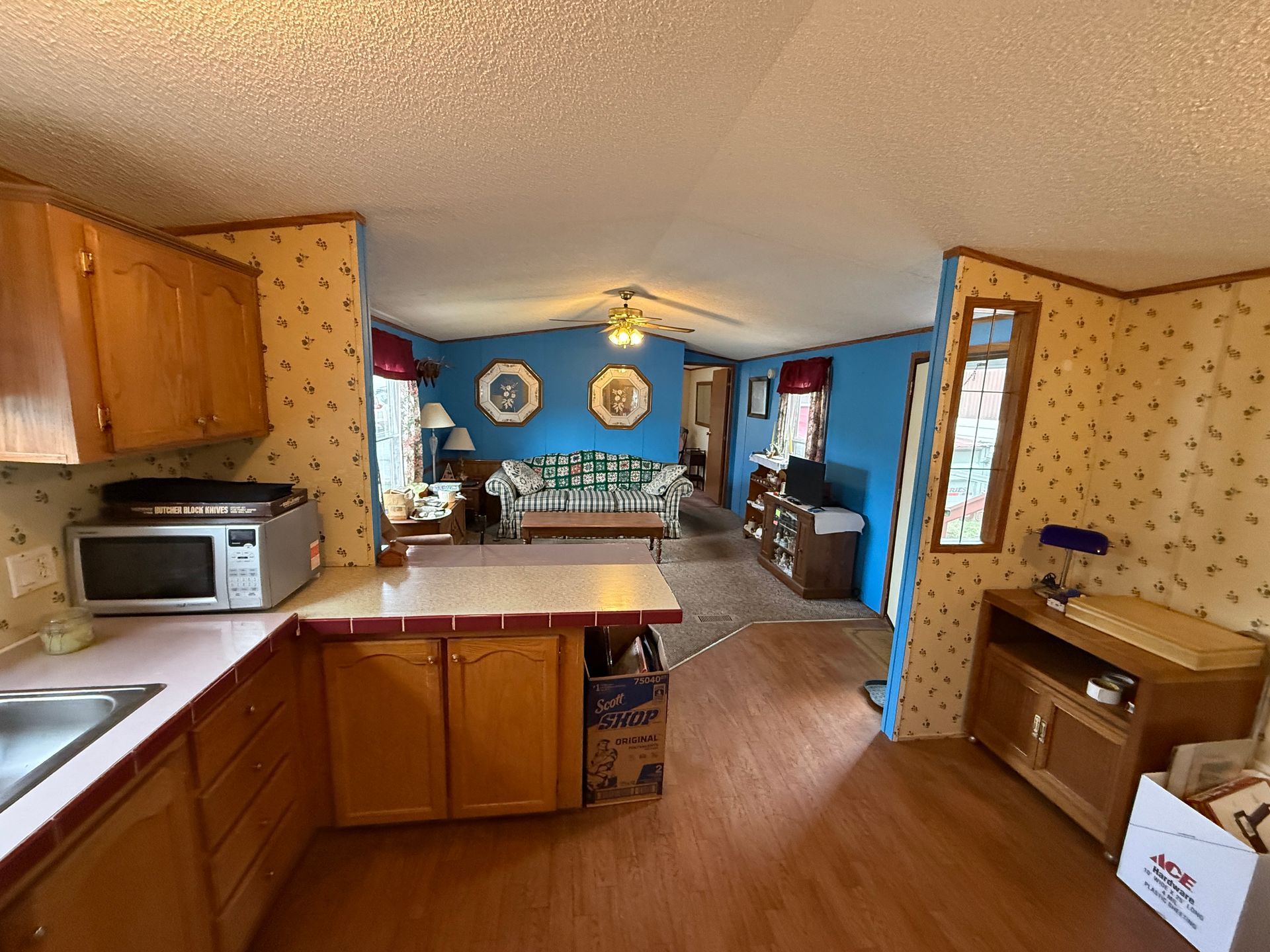 Kitchen with wooden cabinets opening into a blue dining room and living area, with patterned wallpaper and wood floors