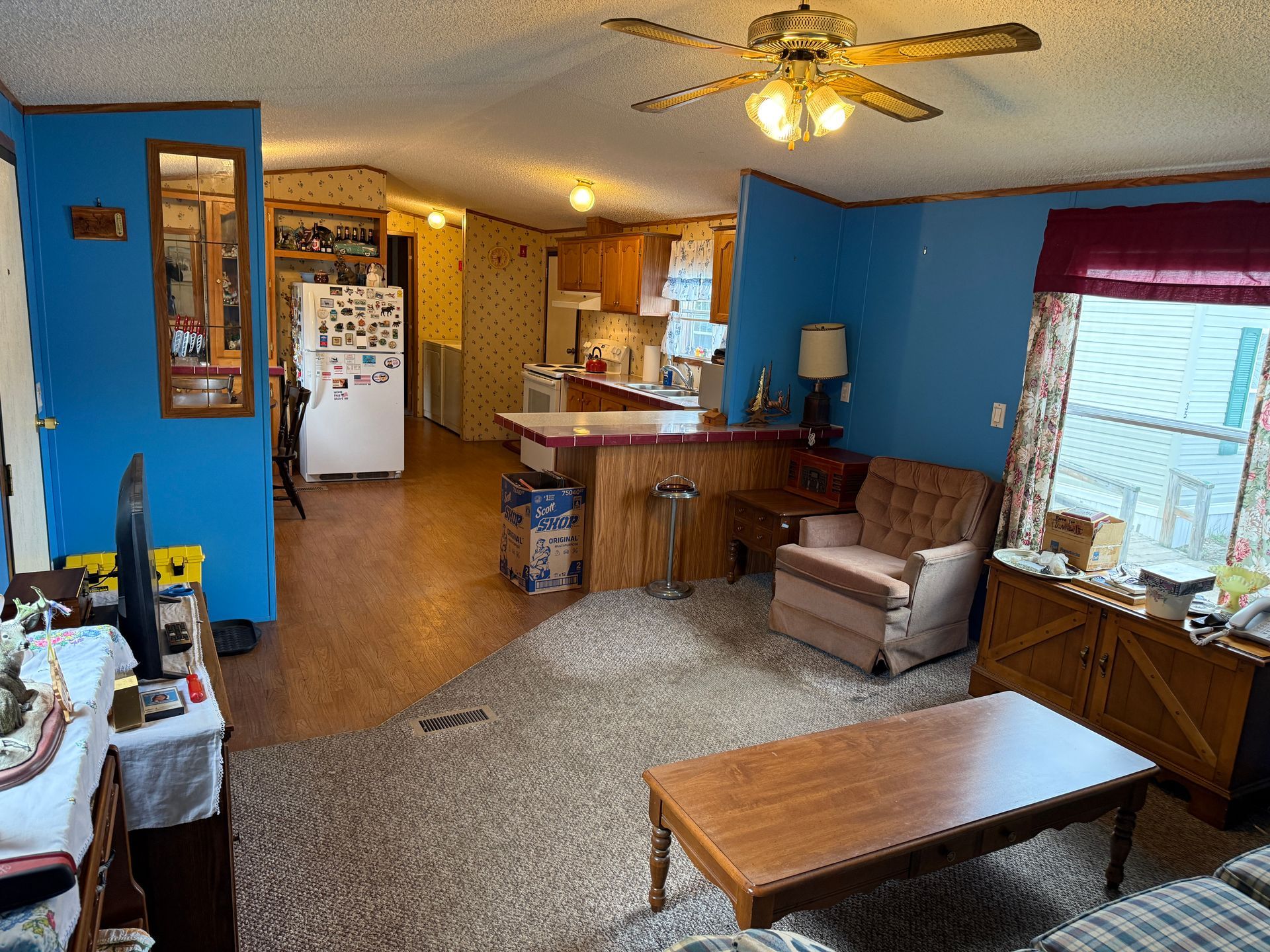 Living room with blue walls, ceiling fan, armchairs, coffee table, and open kitchen in the background